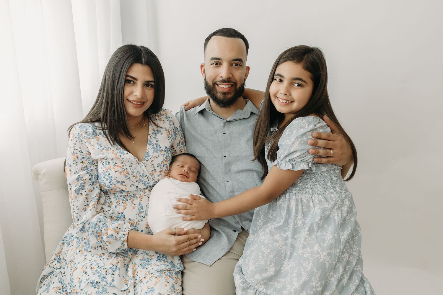 Family in light and airy room wearing coordinated white and gray outfits poses together with newborn baby.