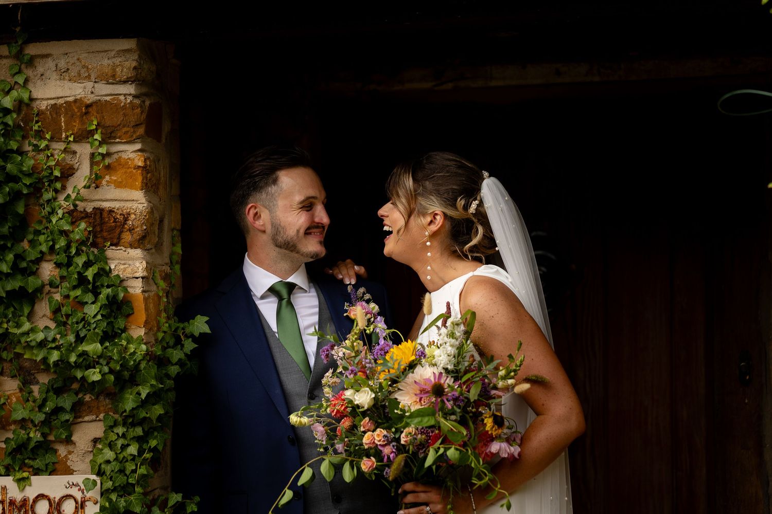 Newlywed couple in navy suit and white dress share a loving moment with colorful wedding bouquet by brick archway.