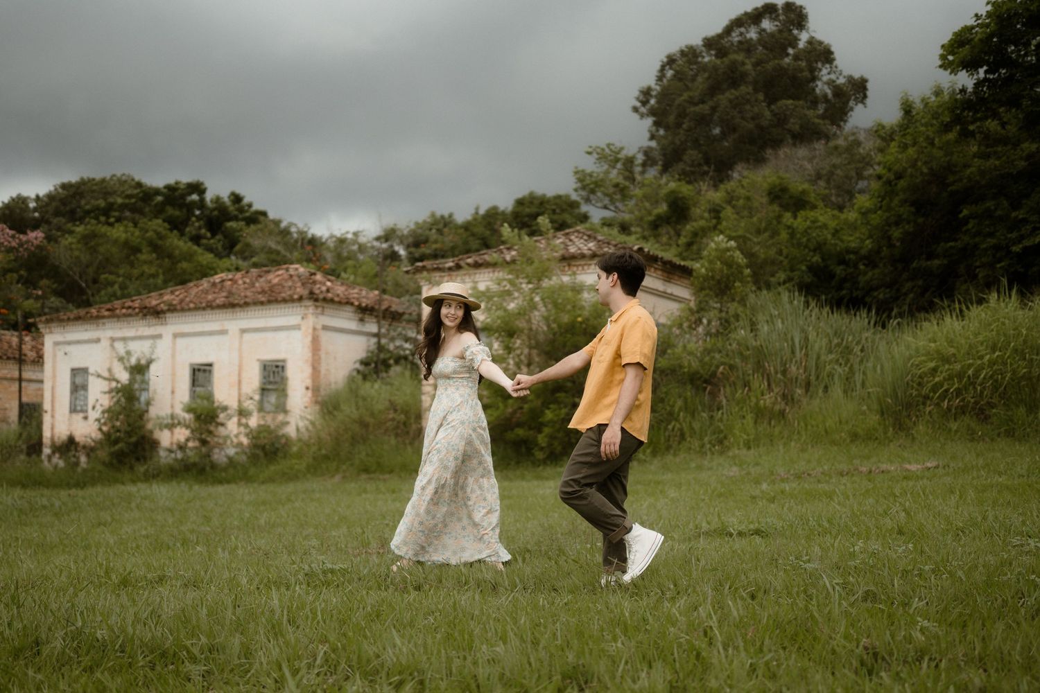Couple in romantic attire walks playfully through green field near abandoned building.