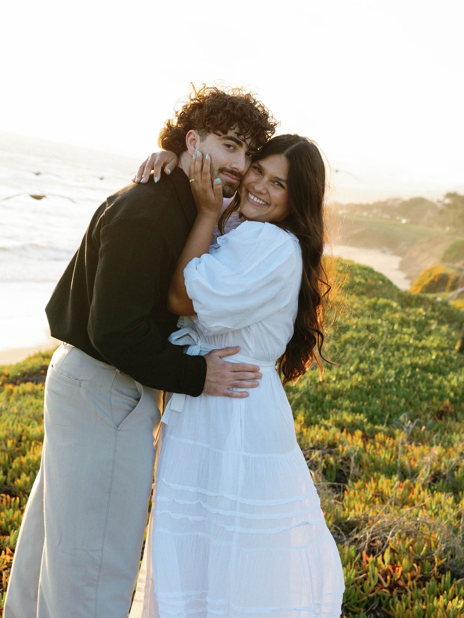 Couple embraces affectionately on a grassy coastal cliff during golden hour, wearing casual white and black outfits.