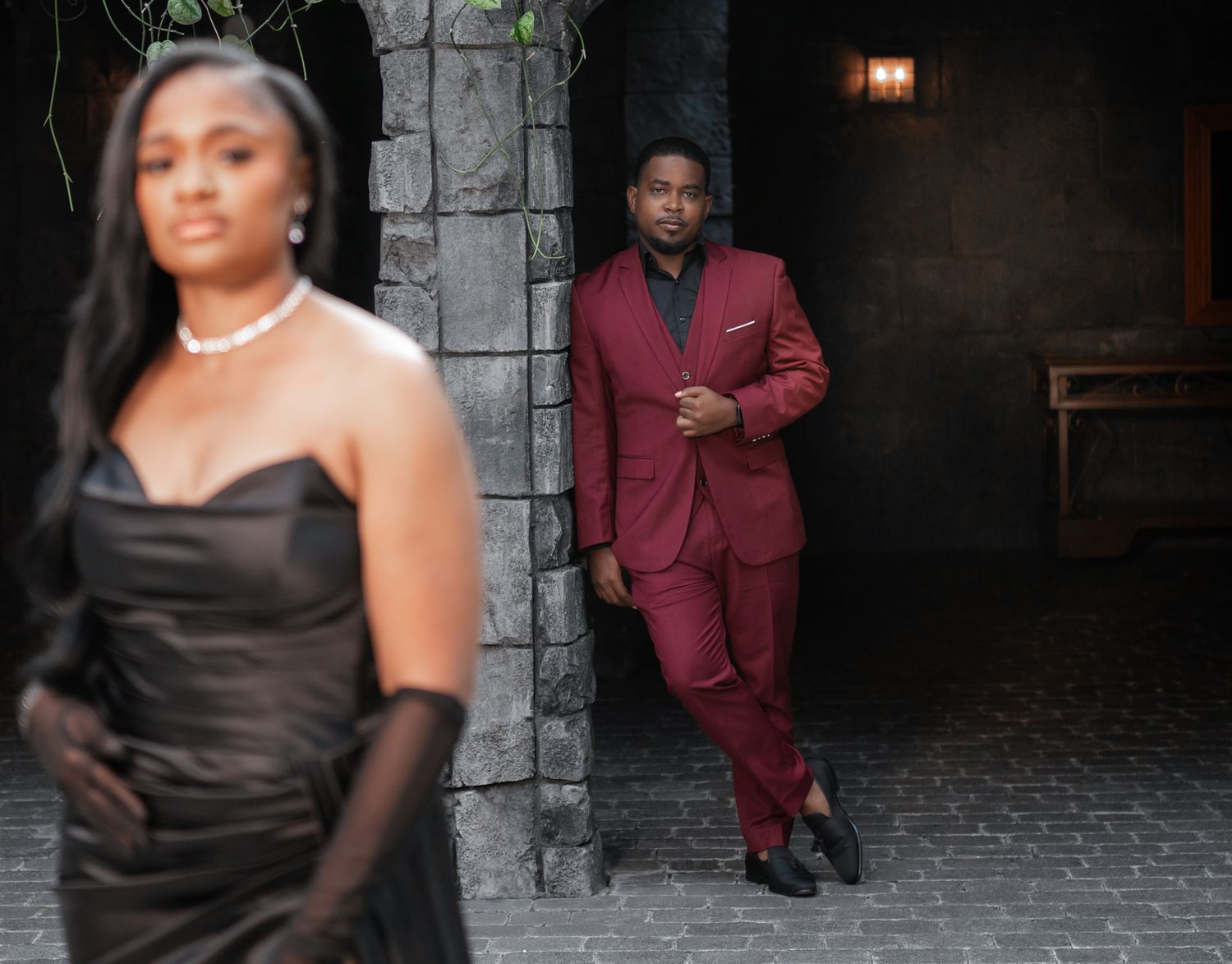 A woman in a black satin gown poses confidently as a man in a burgundy suit stands in the background under stone arch in romantic setting.