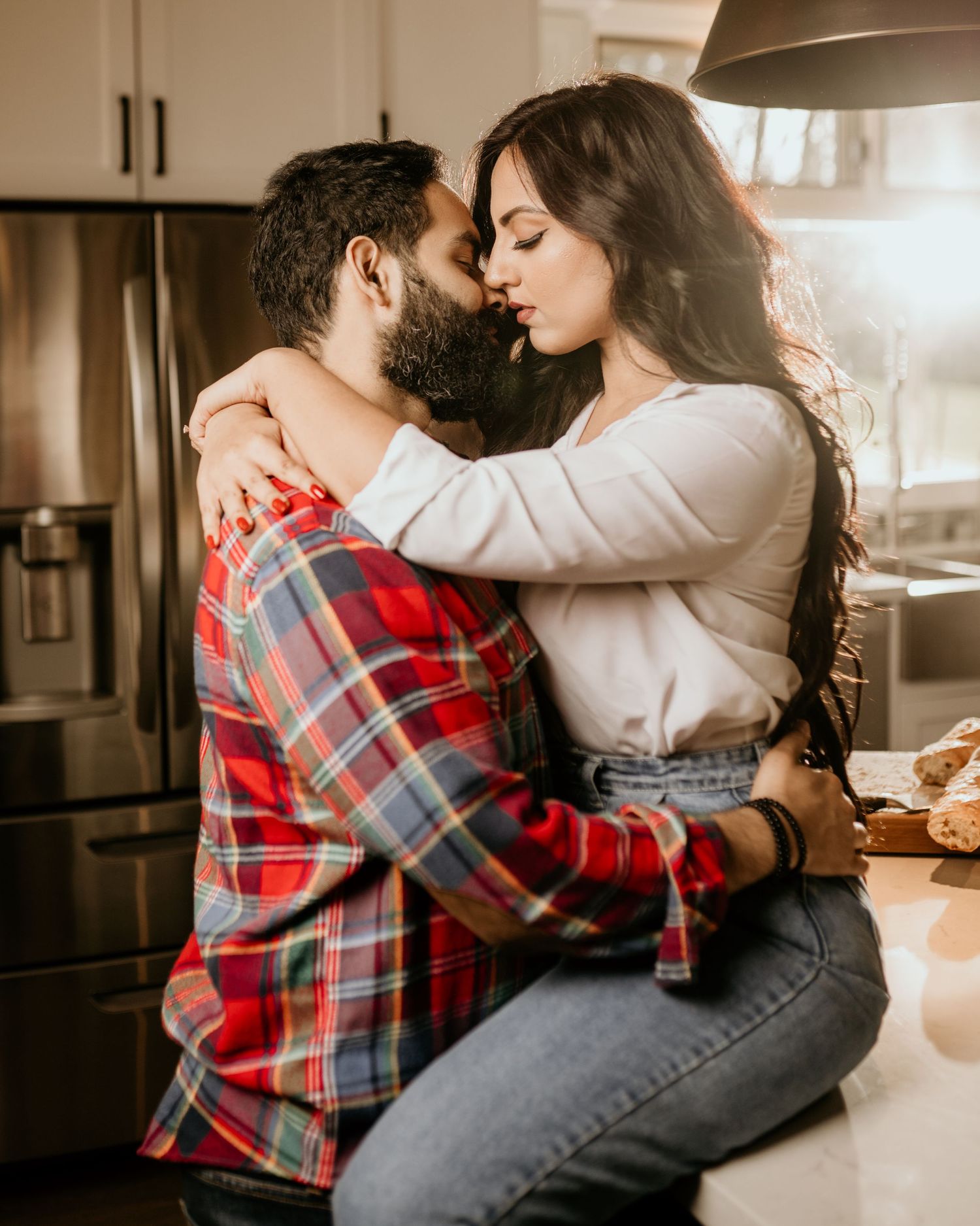 A romantic couple embraces intimately in their kitchen, with warm sunlight streaming through the window.