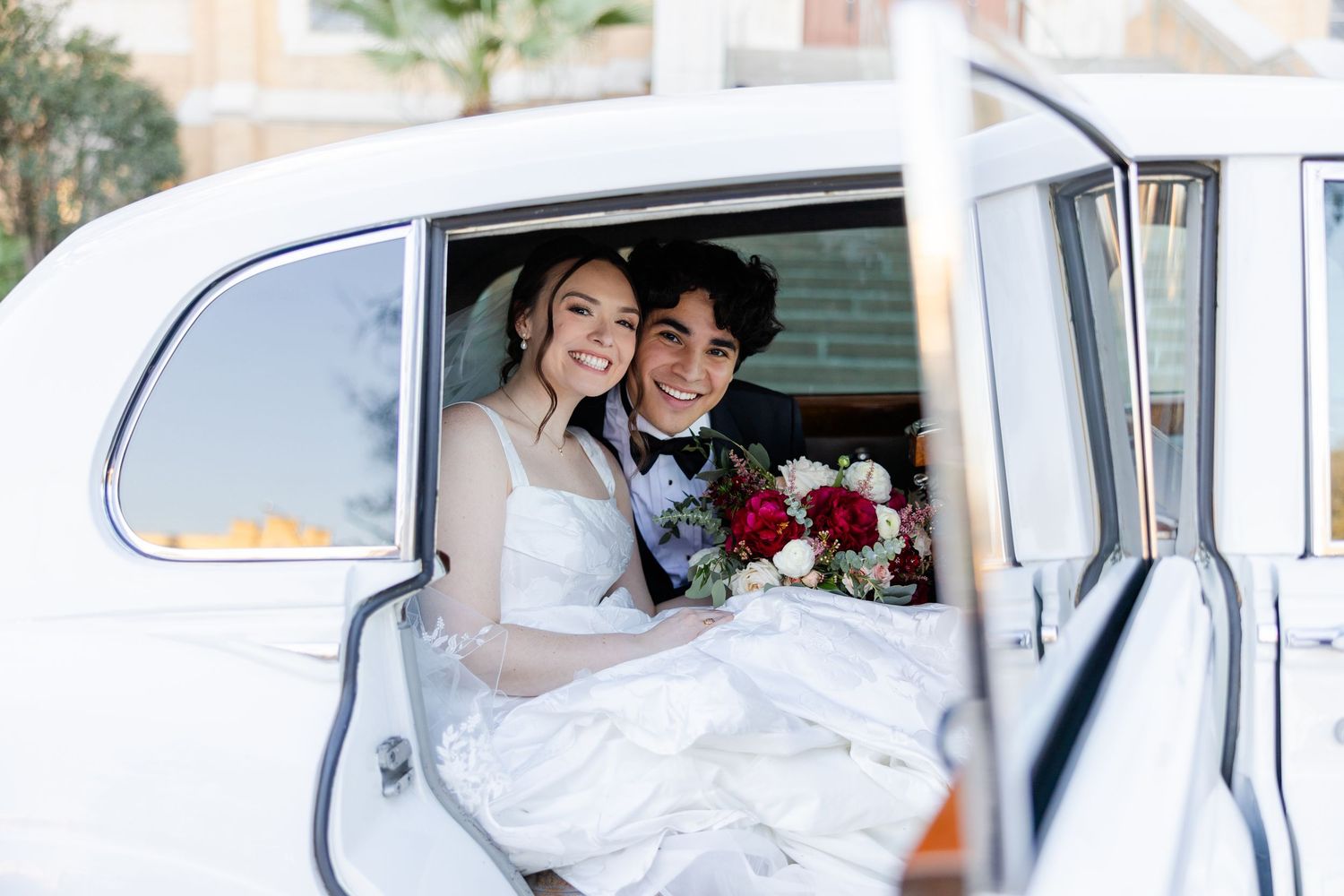 Happy newlyweds smile while sitting in a classic white vintage car with red bouquet.