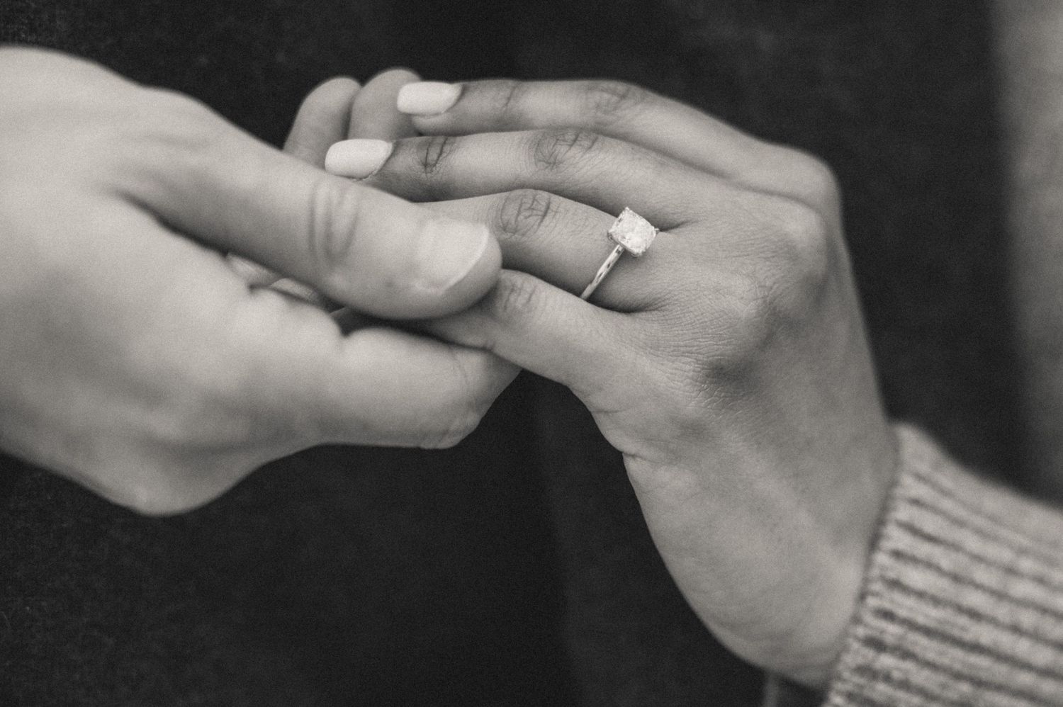A close-up black-and-white photo of a man’s and woman’s hands with an engagement ring during a secret proposal.