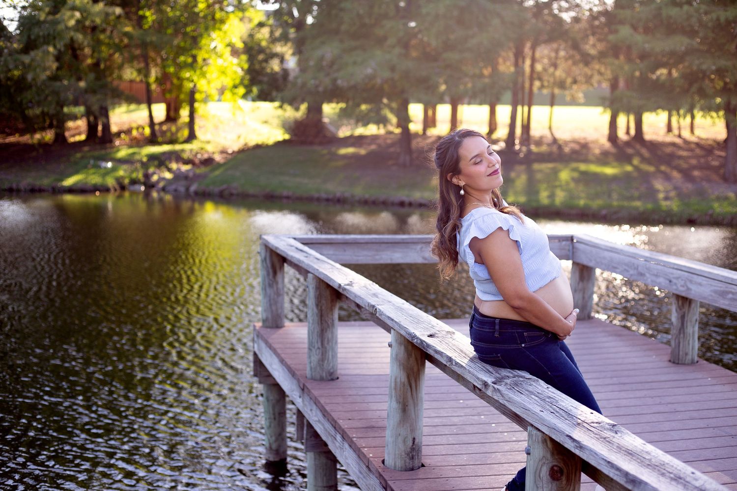 A pregnant woman gazes at the water while standing on a wooden dock at sunset.