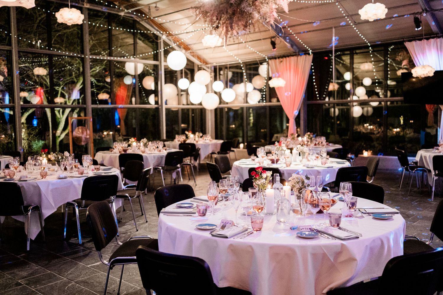 An elegant indoor reception venue with round tables set with white linens and glowing pendant lights overhead.