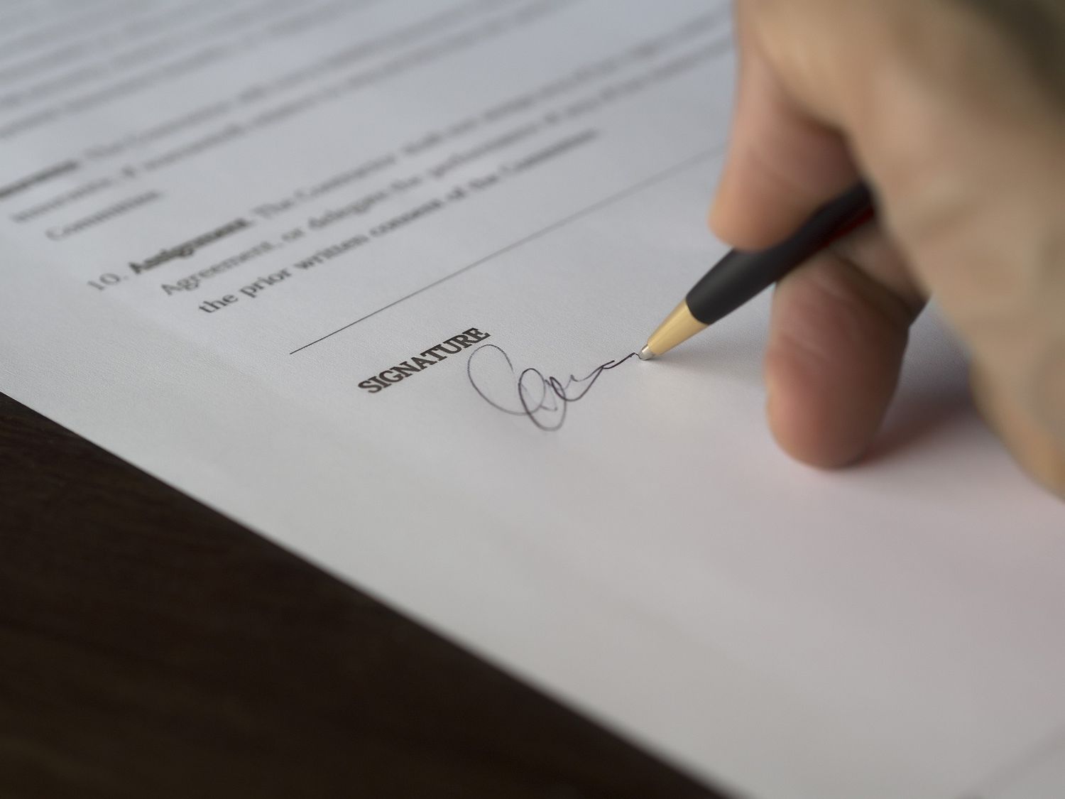 A hand holding a pen signs a signature on an official document against a white paper background.