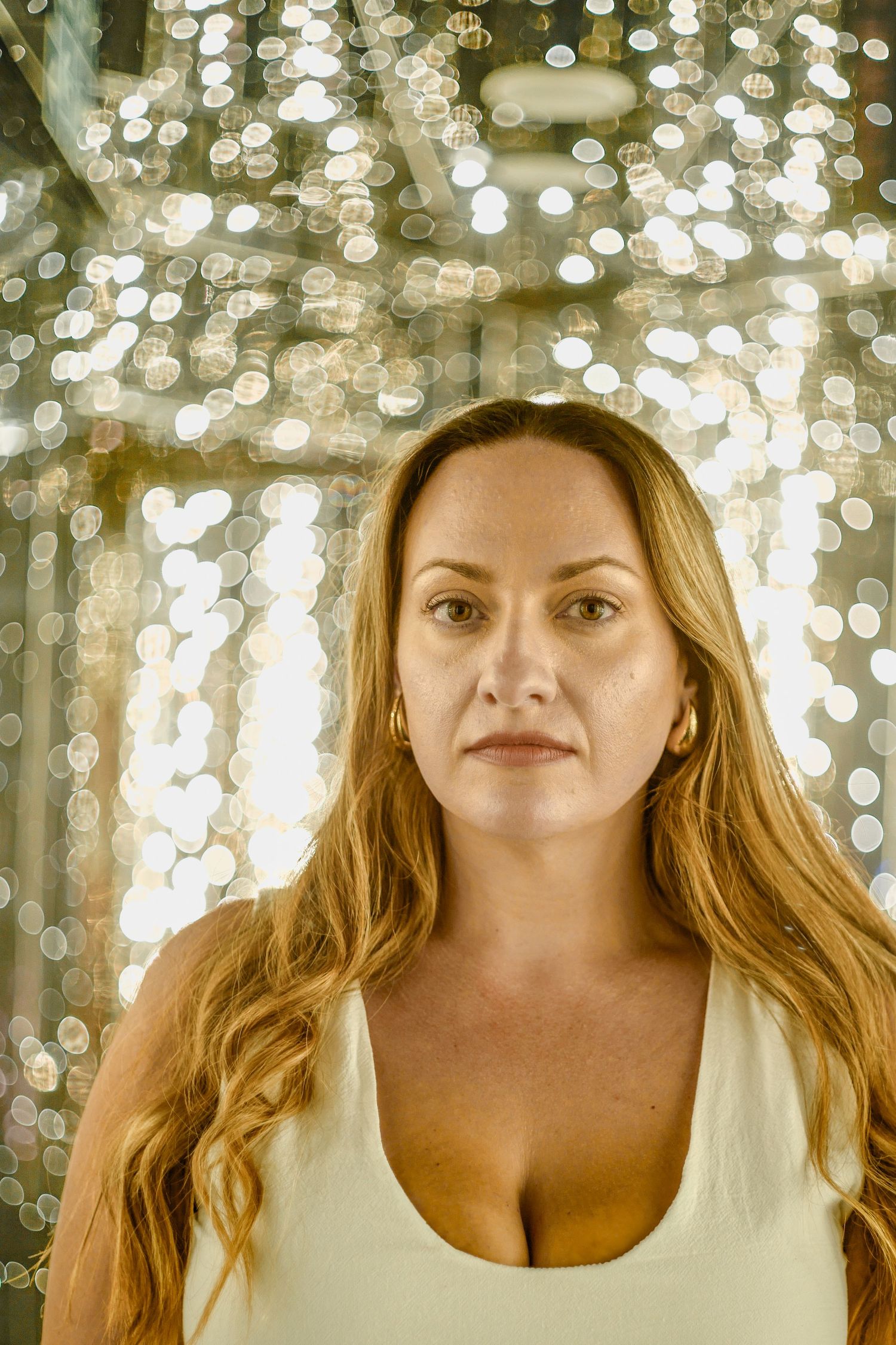 Portrait of someone with long blonde hair wearing a white tank top against a bokeh light background of golden sparkles.
