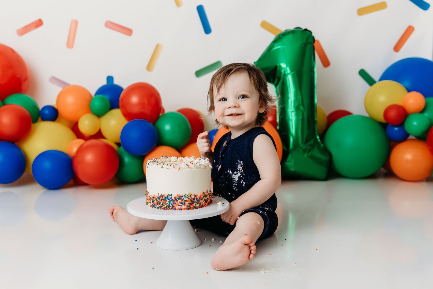 Colorful first birthday party setup with cake on white stand, balloons, and sprinkle decorations against white backdrop.