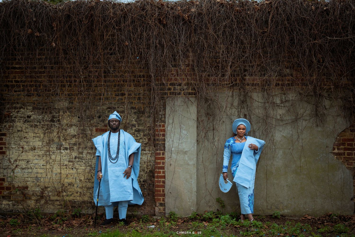 Two people in matching light blue traditional African attire stand against a brick wall covered in vines.