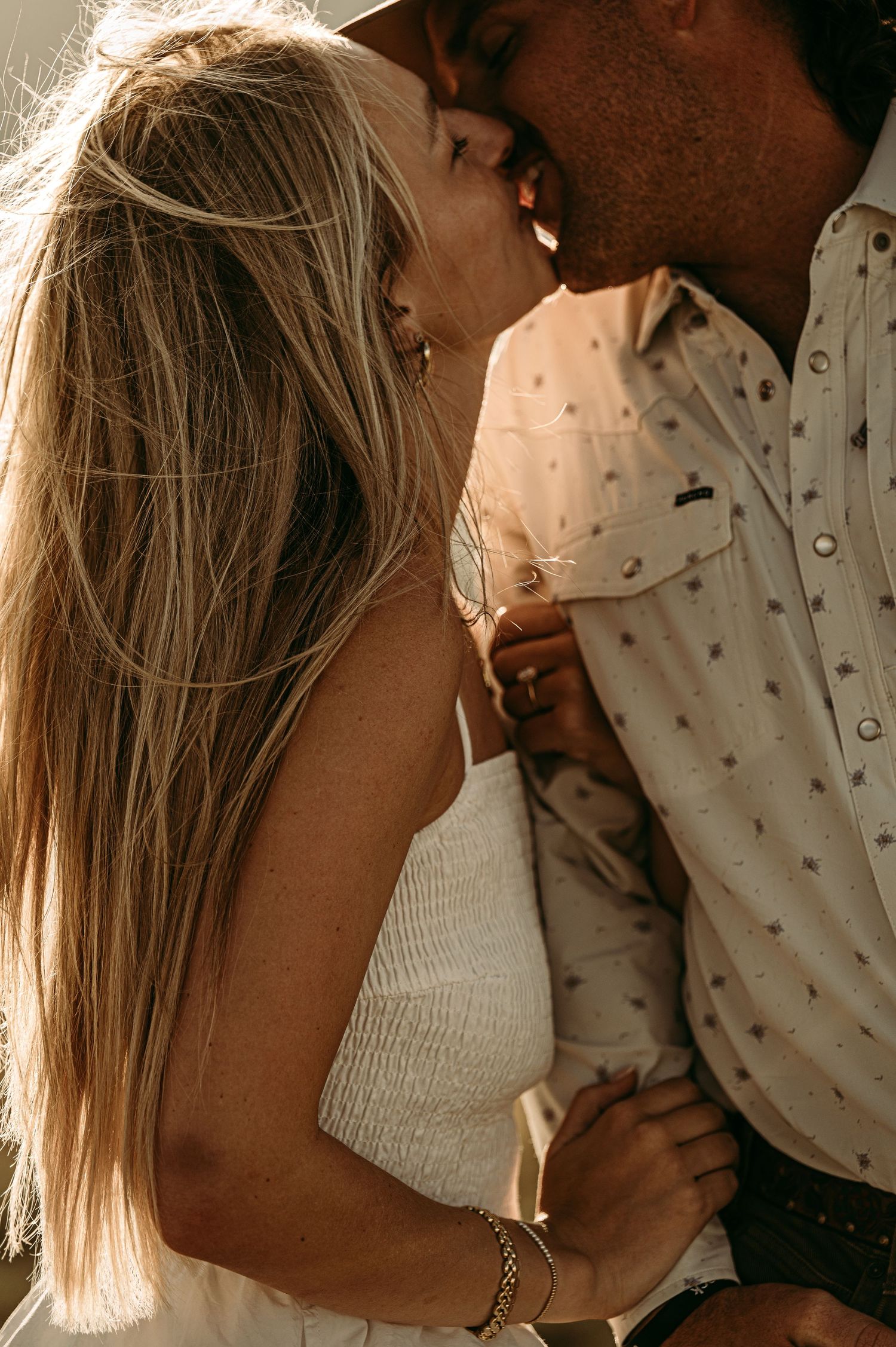 A couple sharing a romantic kiss outdoors, woman with long blonde hair in white dress, man in patterned shirt.