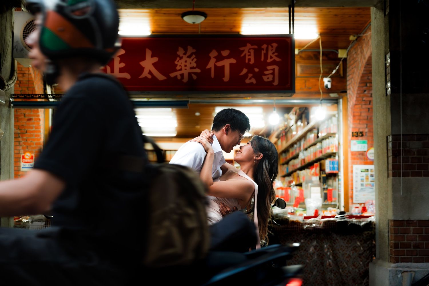 Couple sharing a romantic kiss outside a traditional Chinese pharmacy shop at night with motorcycles parked nearby.