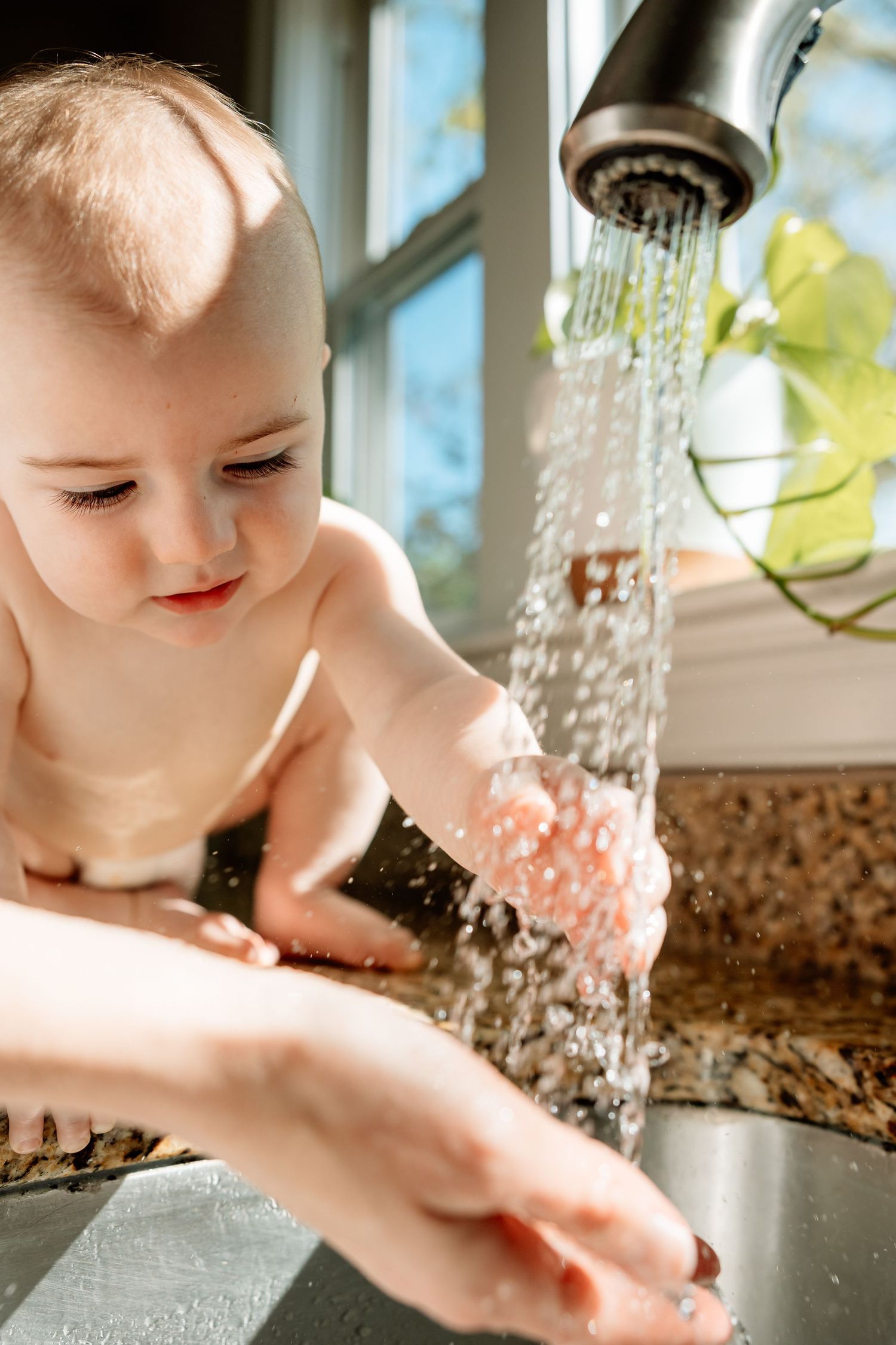 A baby reaches hands under running kitchen faucet water, assisted by an adult, in a bright sunlit home.