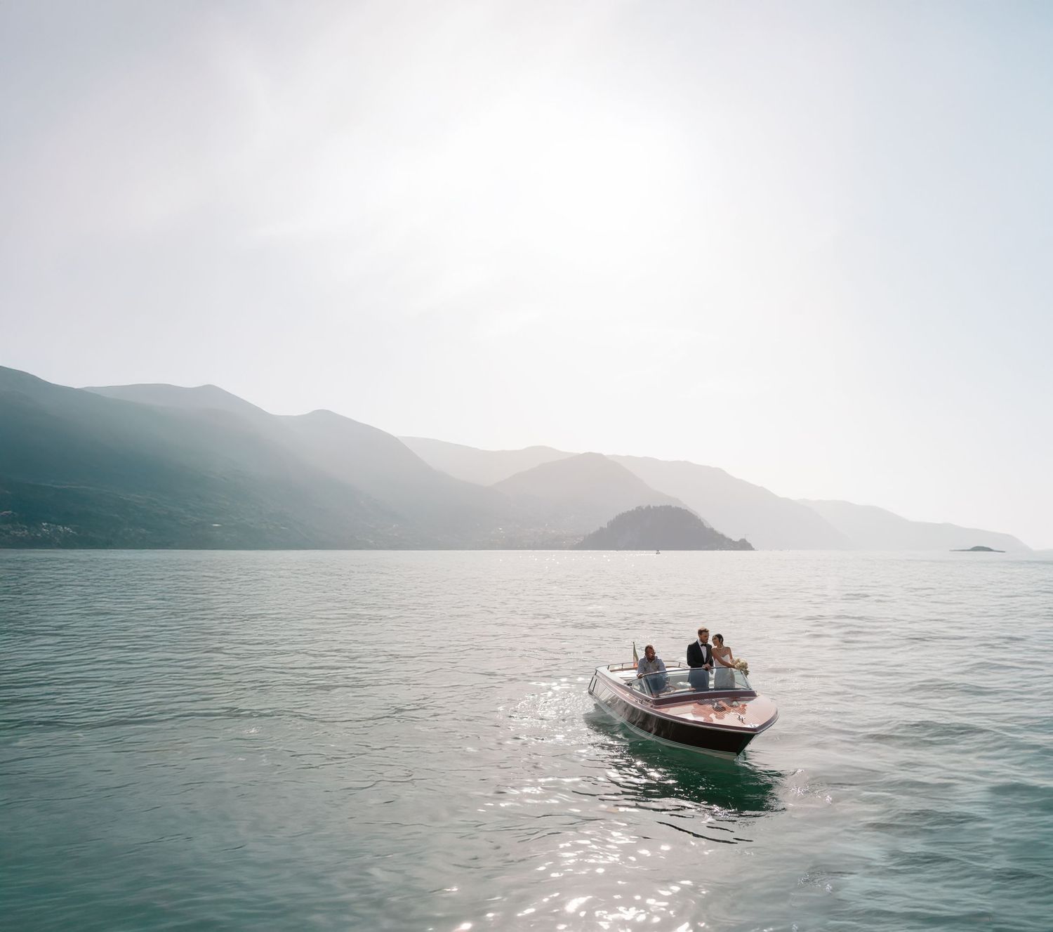 Small motorboat cruises across misty mountain lake on a hazy morning with sunlight reflecting off calm waters.