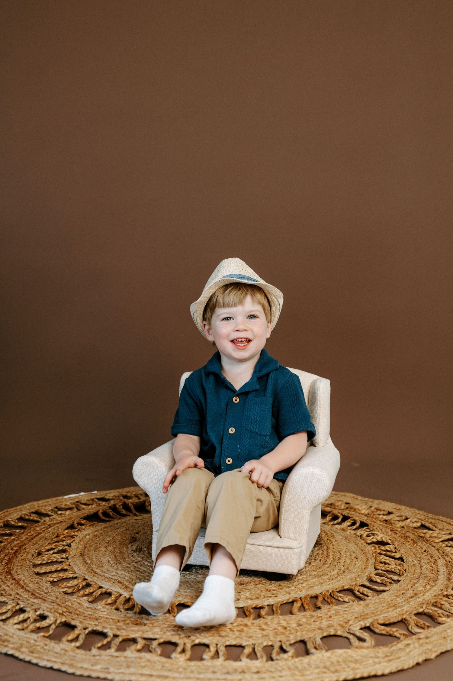 Smiling young boy in a tan hat, navy shirt, and khaki pants sitting in a small chair on a woven rug.