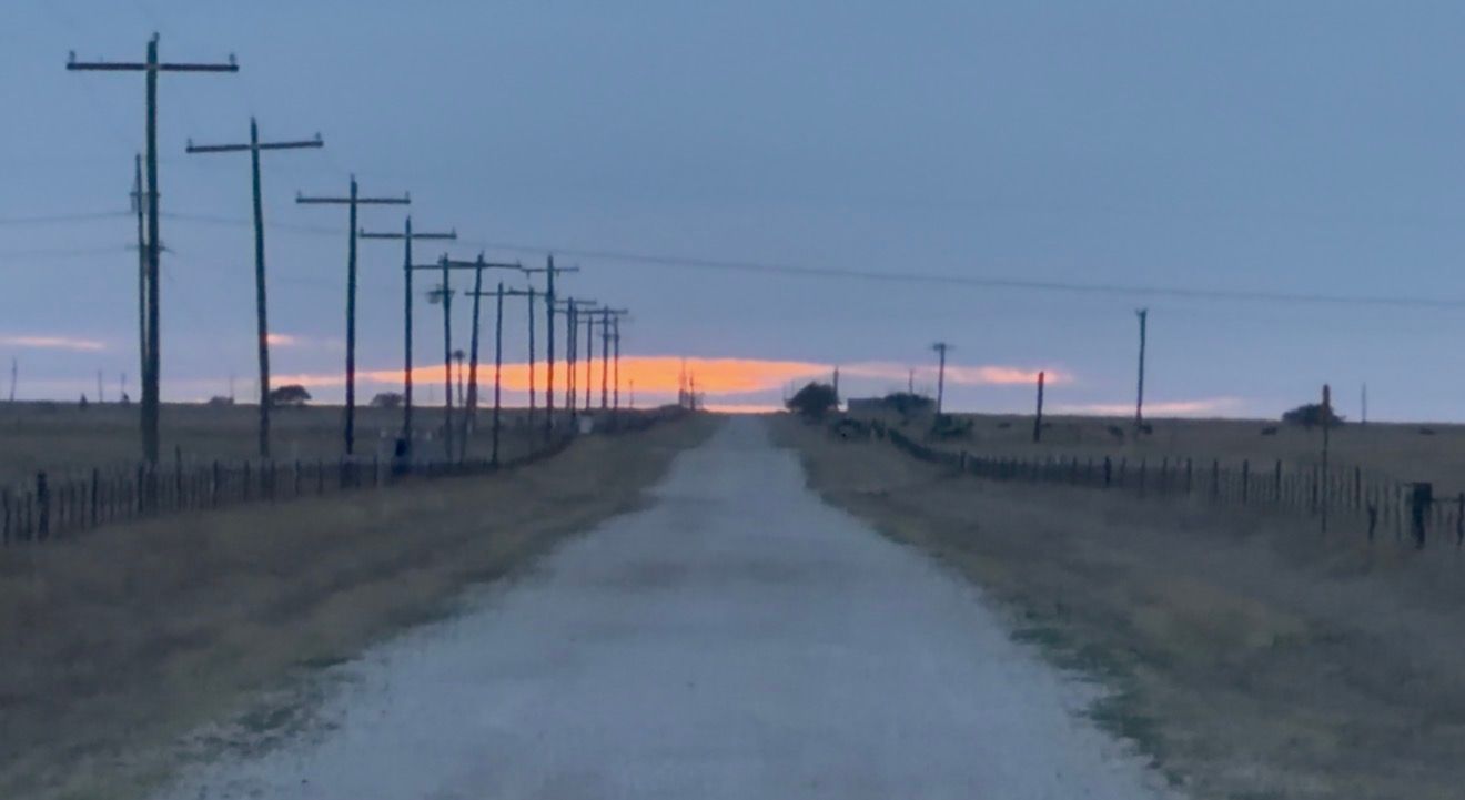 Rural road at dusk with telephone poles lining both sides, leading to a vivid orange and blue sunset horizon.