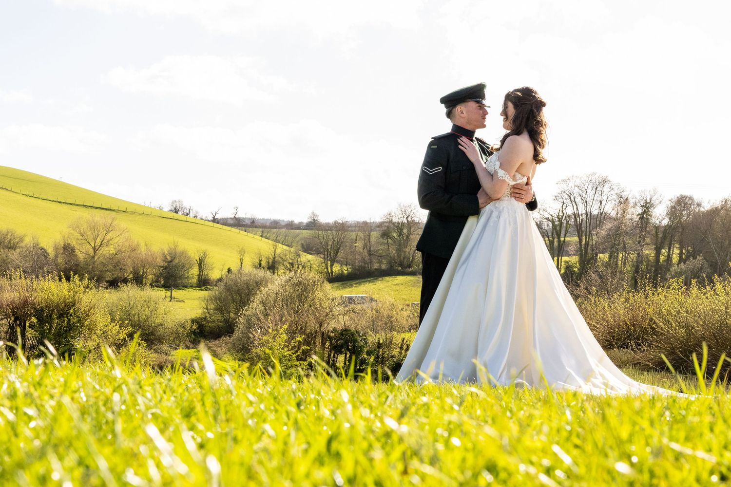 Bride and groom embracing in a sunlit green field, with rolling hills in the background on their wedding day.