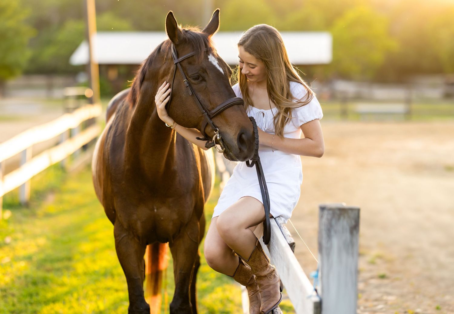 Young woman in white dress and brown boots smiling at a dark brown horse in a sunny ranch setting.