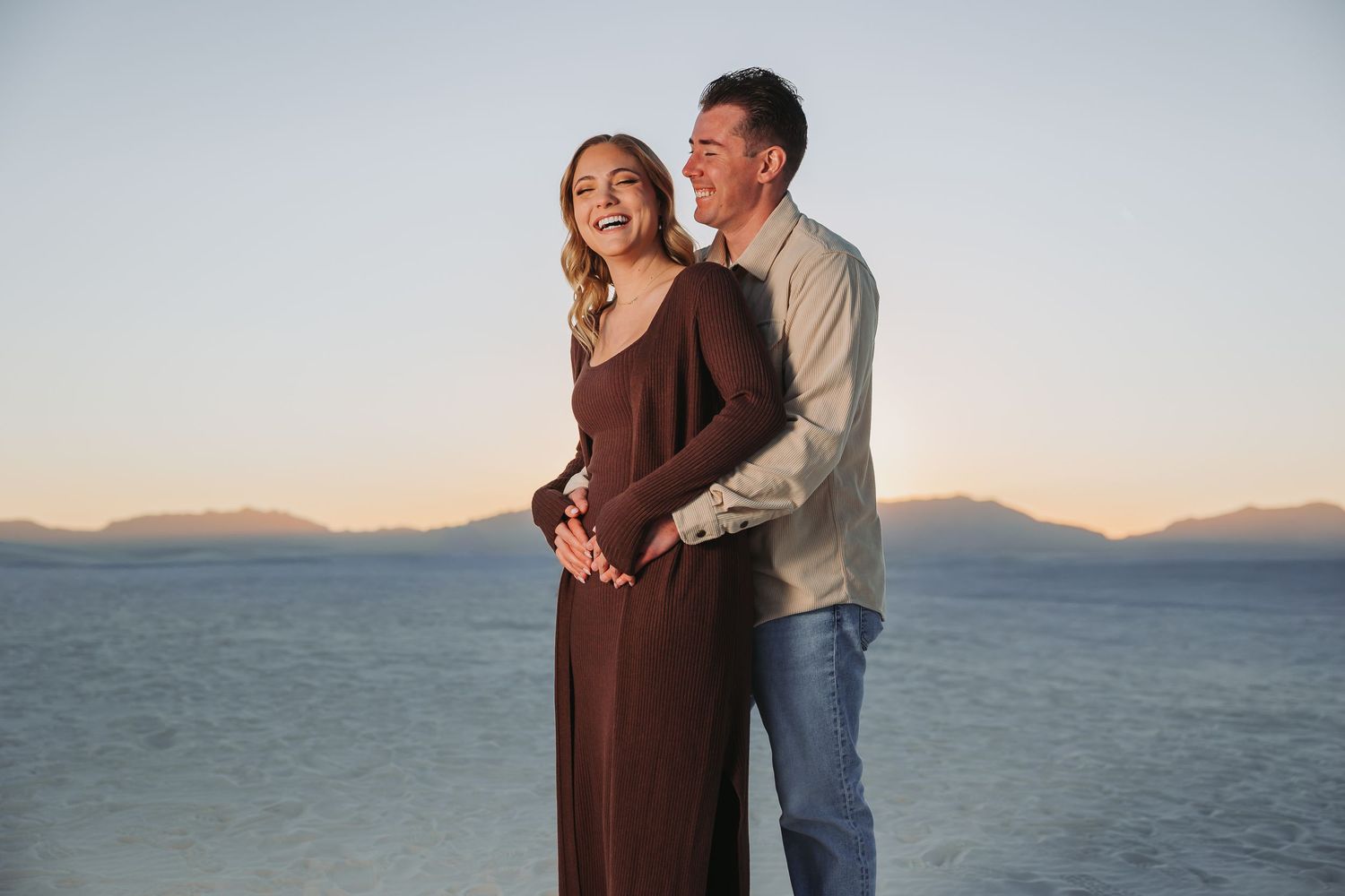 Couple embraces while standing on beach at sunset with mountains in background.