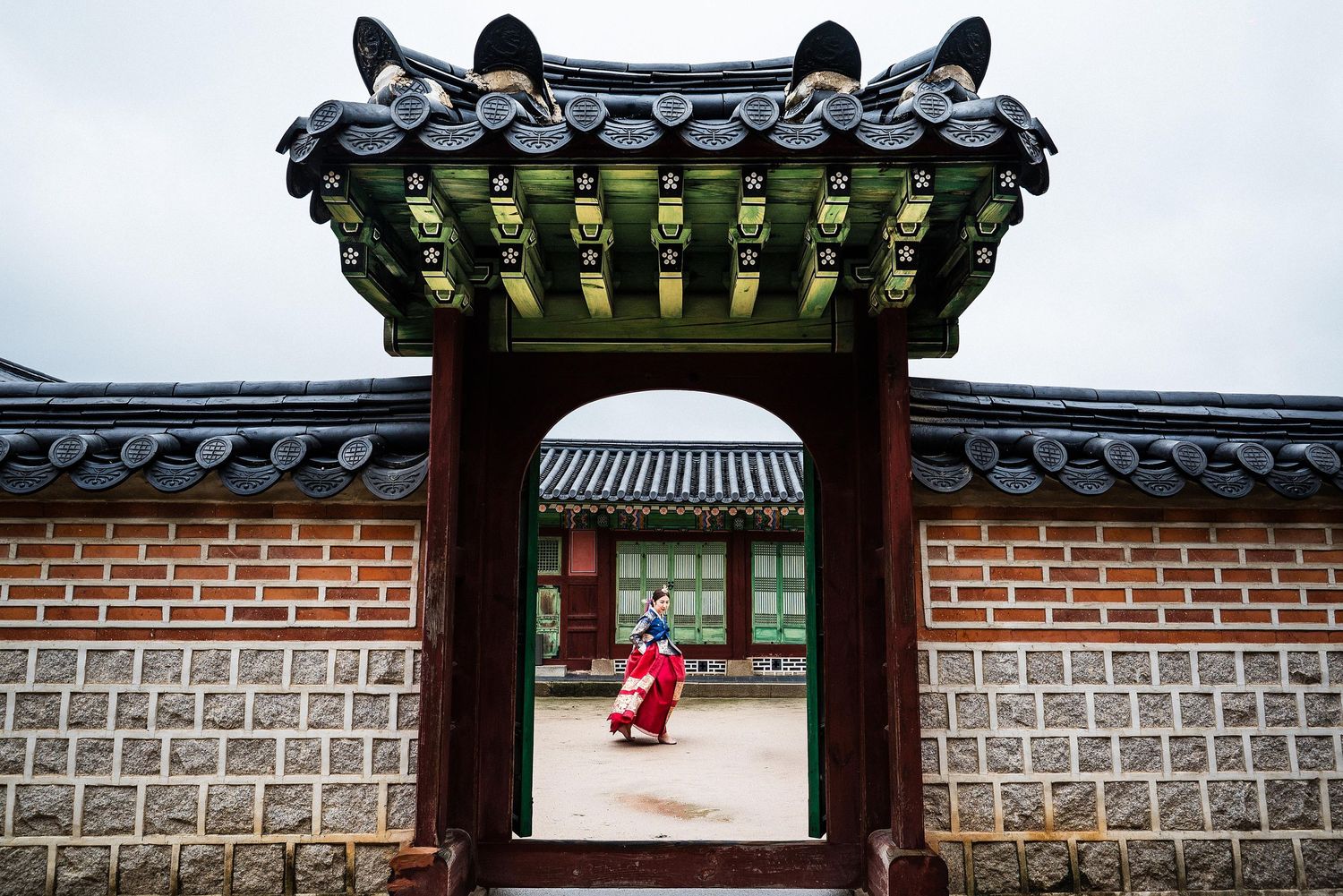 Person in traditional Korean hanbok visible through ornate palace gate with brick walls and decorative roof.