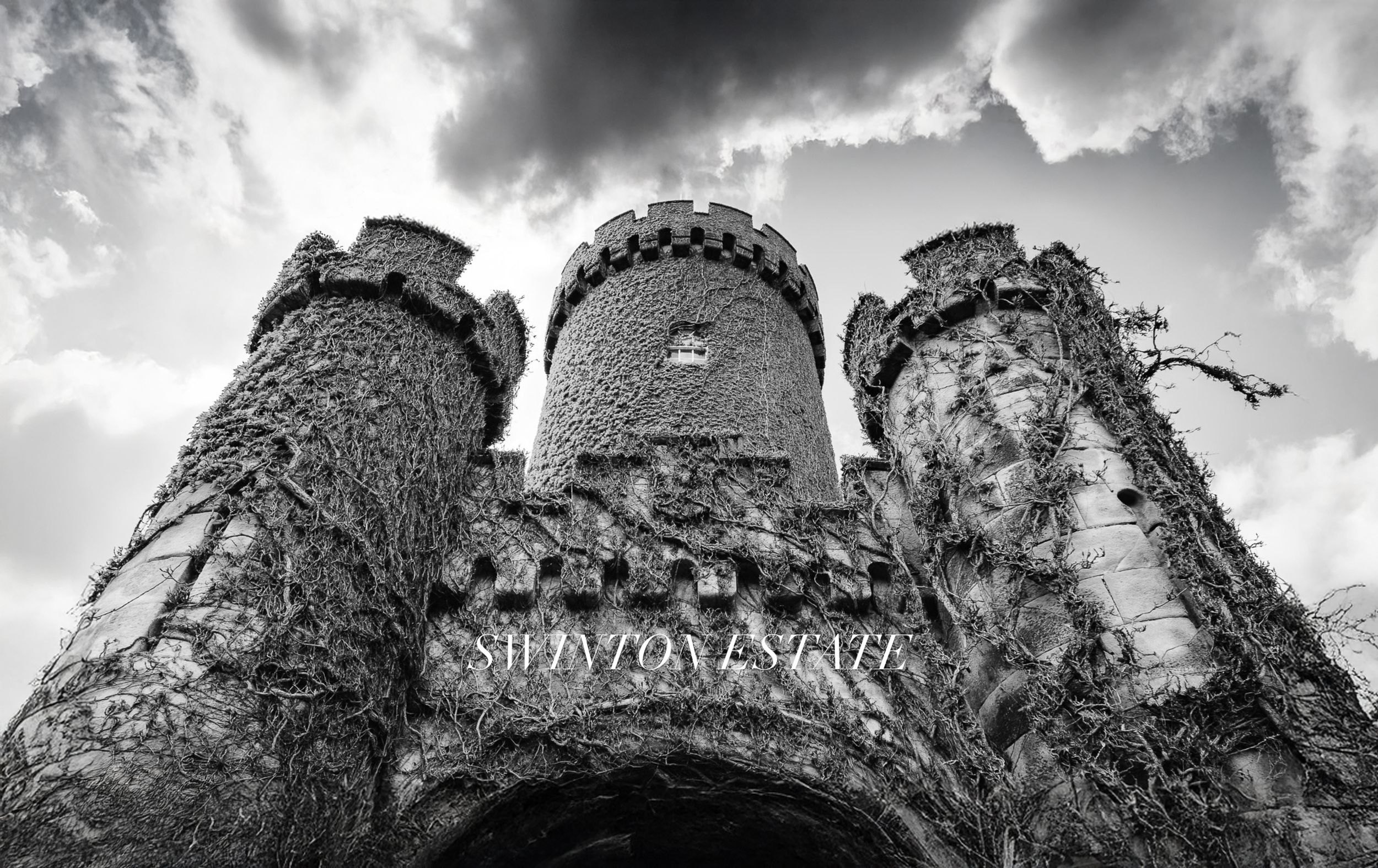 Black and white photo of Swinton Estate castle entrance with ivy-covered stone towers against dramatic cloudy sky.