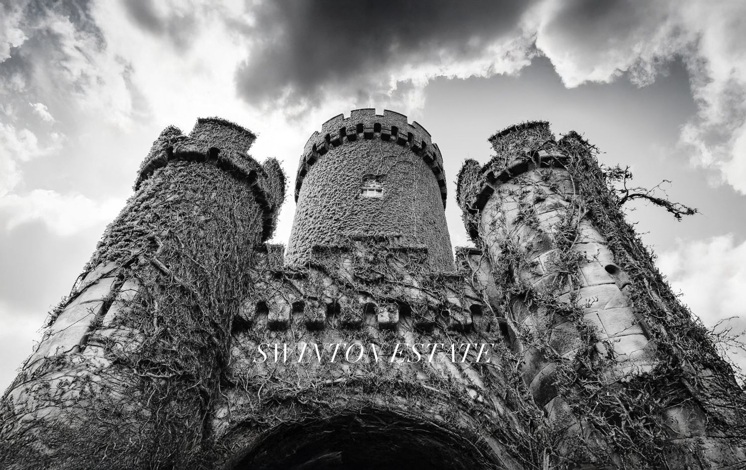 Black and white photo of Swinton Estate castle entrance with ivy-covered stone towers against dramatic cloudy sky.
