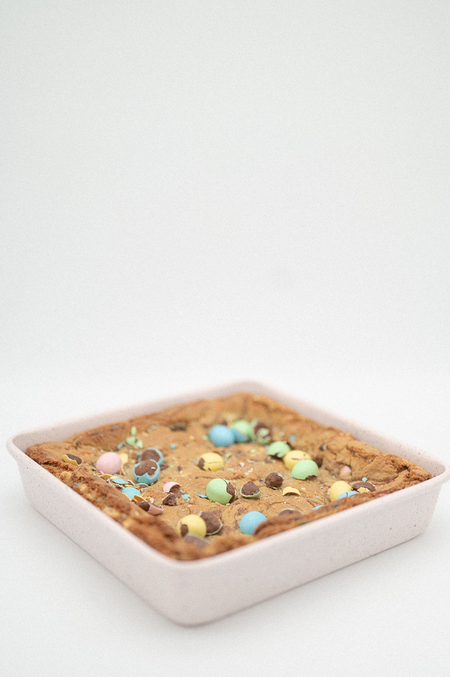 Golden cookie bar with chocolate chips and pastel candy eggs baked in a white square pan.