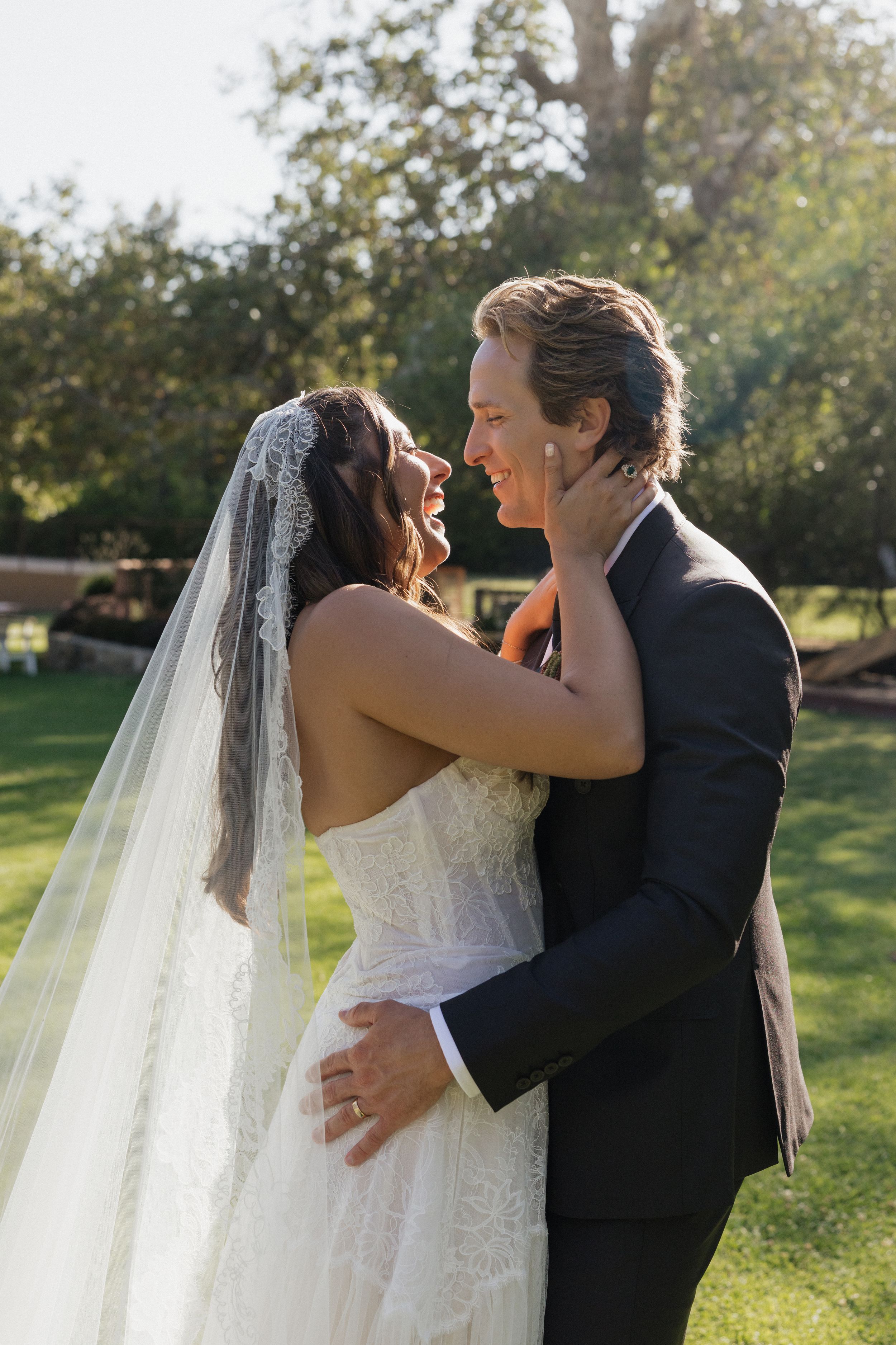 A bride and groom share a joyful moment outdoors, embracing and smiling at each other in wedding attire.