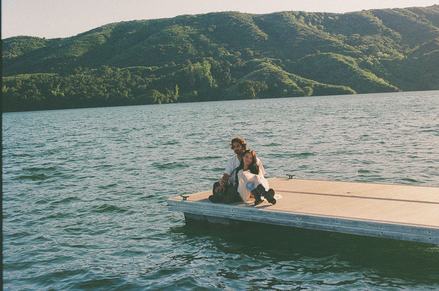 Person sitting on a white dock by a calm lake surrounded by lush green hills.