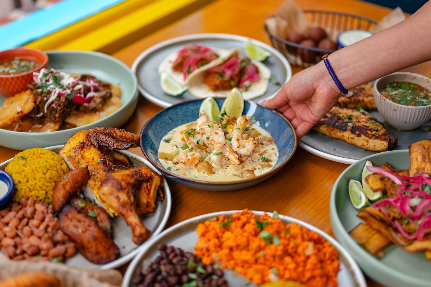 A hand placing lime on a bowl of creamy dip surrounded by colorful Mexican dishes on a wooden table.