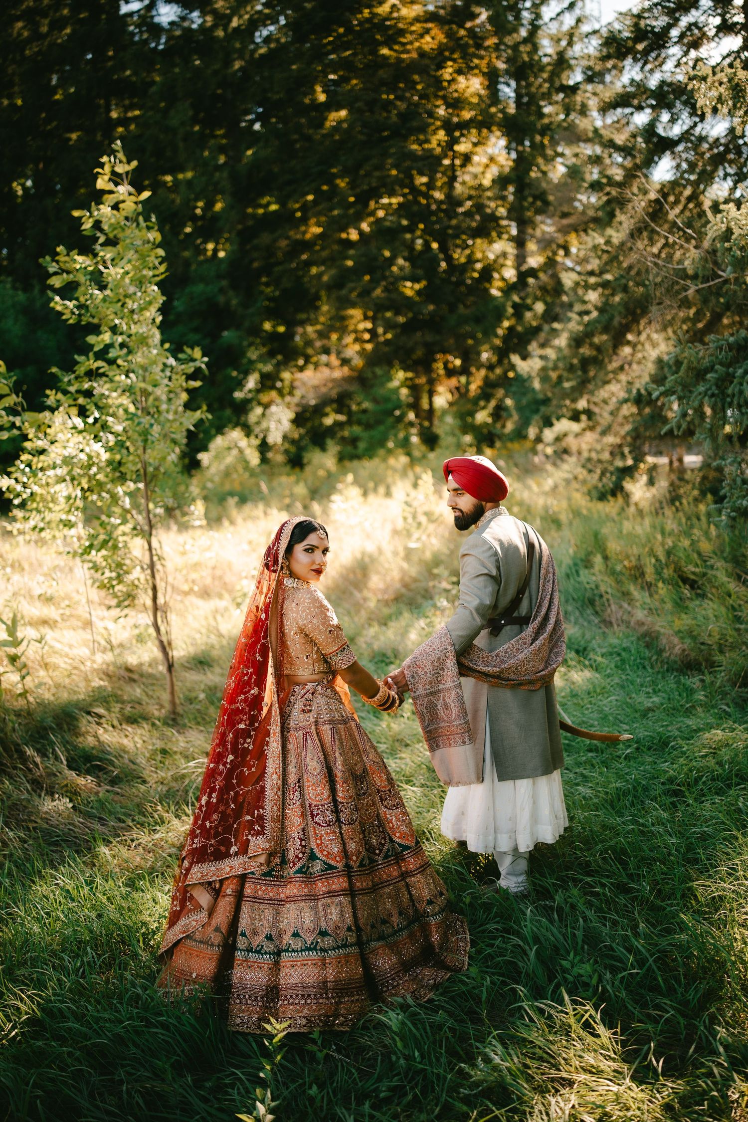 A Sikh bride in red lehenga and groom in gray sherwani with red turban hold hands in a lush green forest setting.