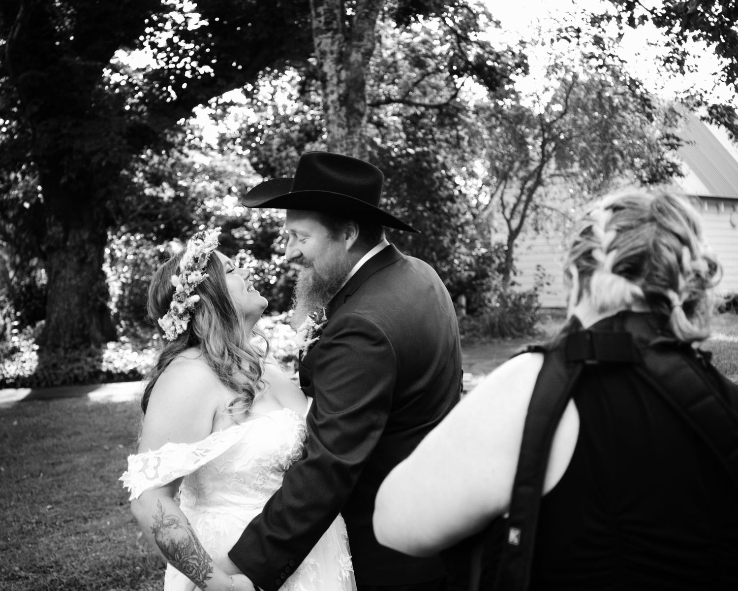 Black and white wedding photo of bride and groom sharing a kiss outdoors, with guest in foreground.