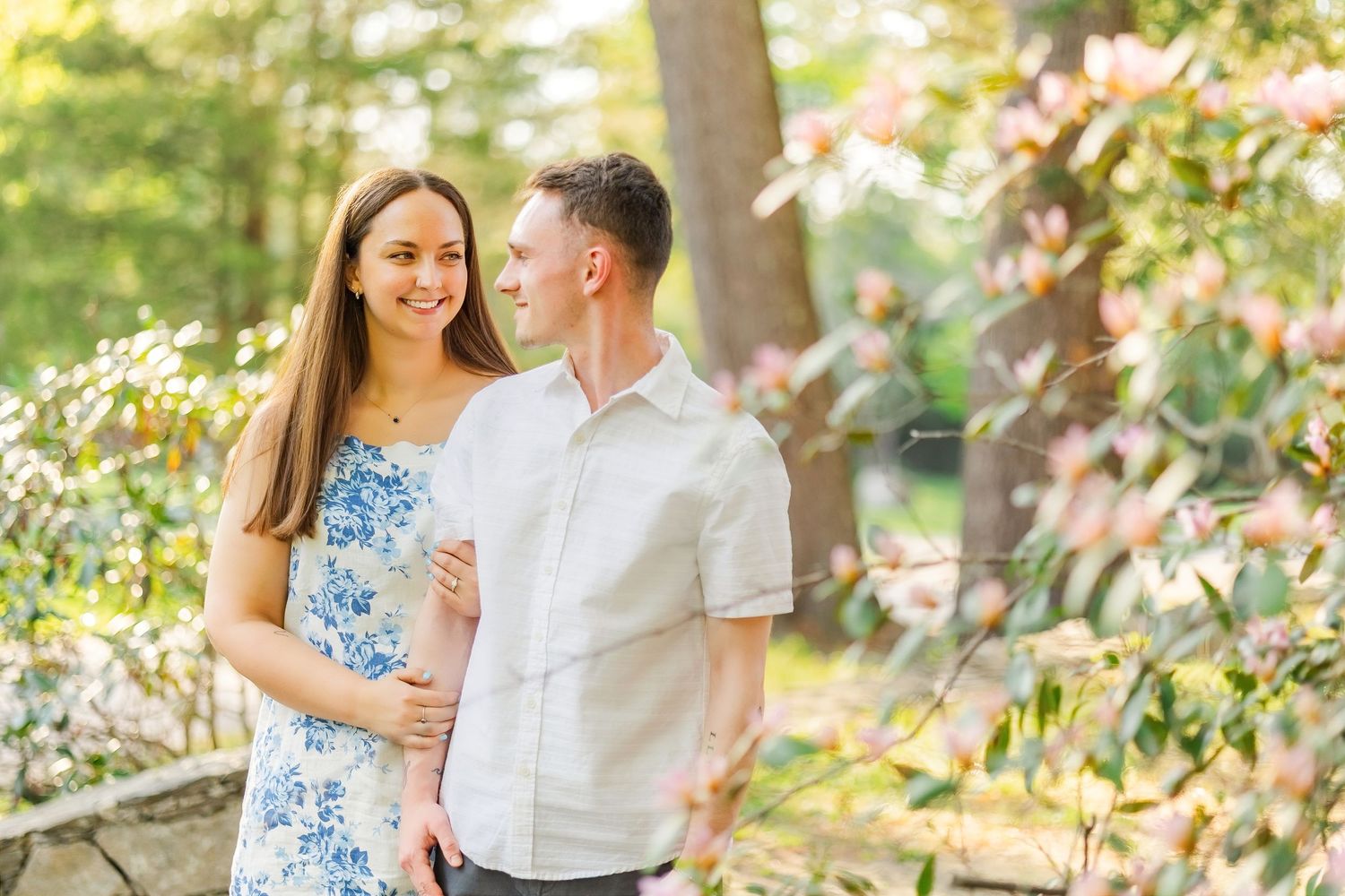 Couple embracing outdoors in spring garden with blooming pink flowers and lush green trees in soft sunlight.