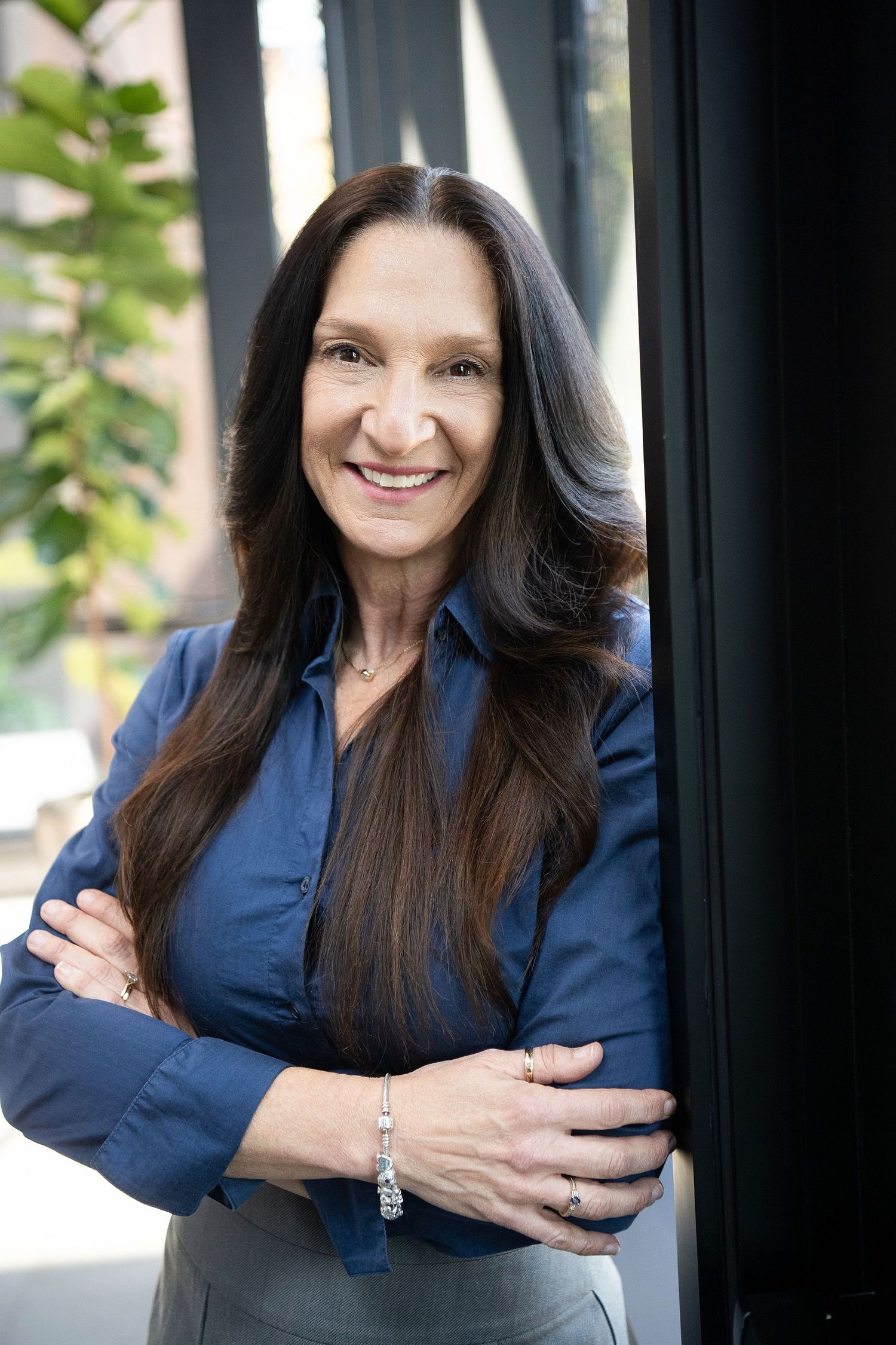 Smiling middle-aged woman with long dark hair in a blue shirt, arms crossed, posing confidently near a window.