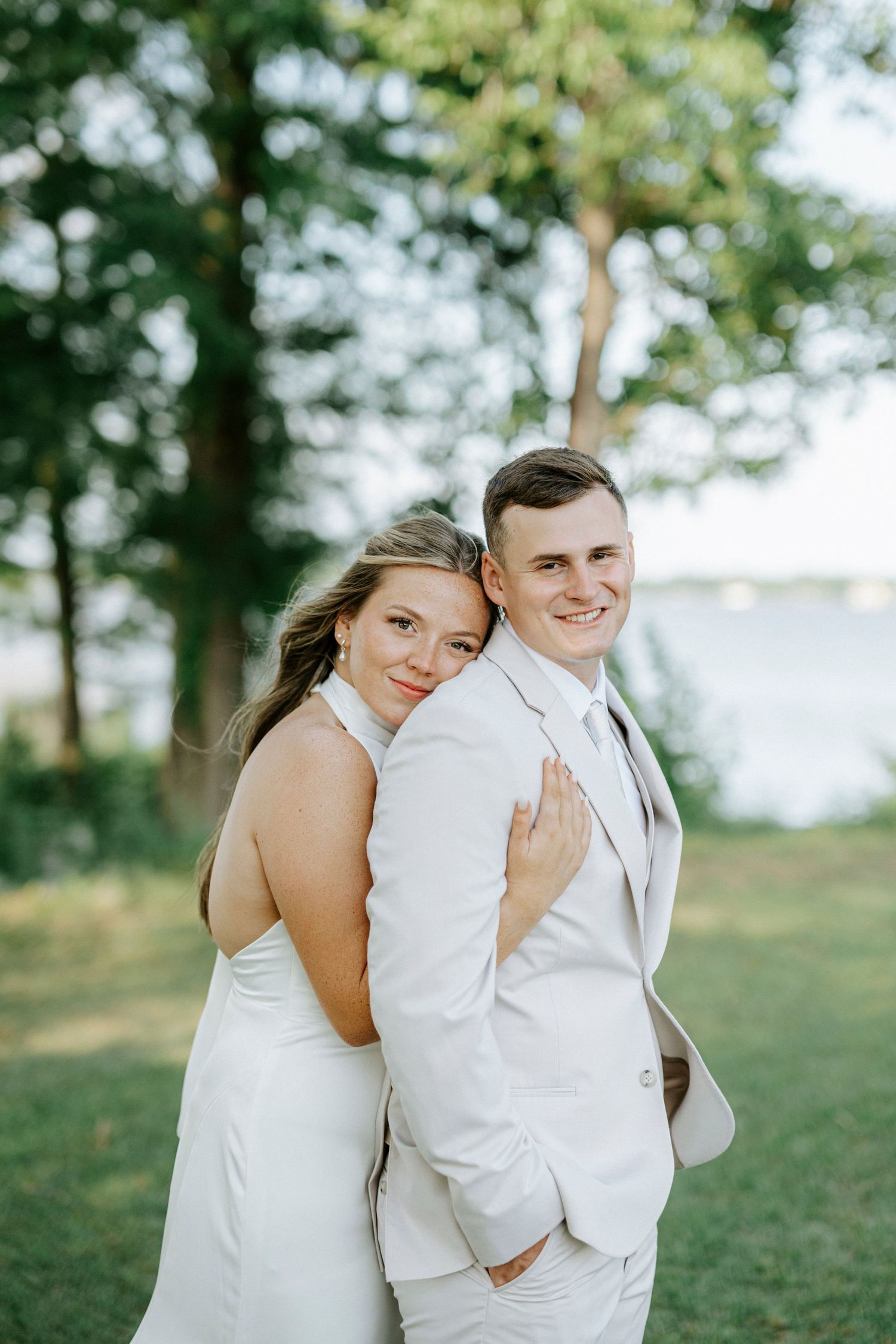 A bride in a white dress embraces a groom in a light suit outdoors near a lake with lush green trees.