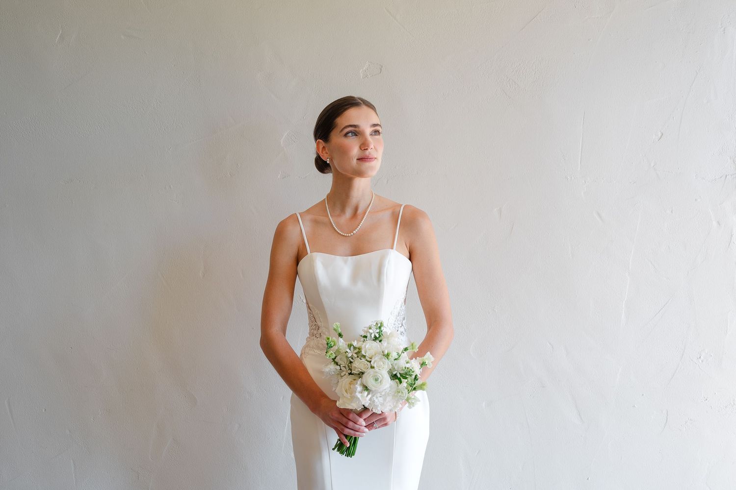 A bride in a sleeveless white wedding dress holds a delicate bouquet of white flowers against a light gray background.