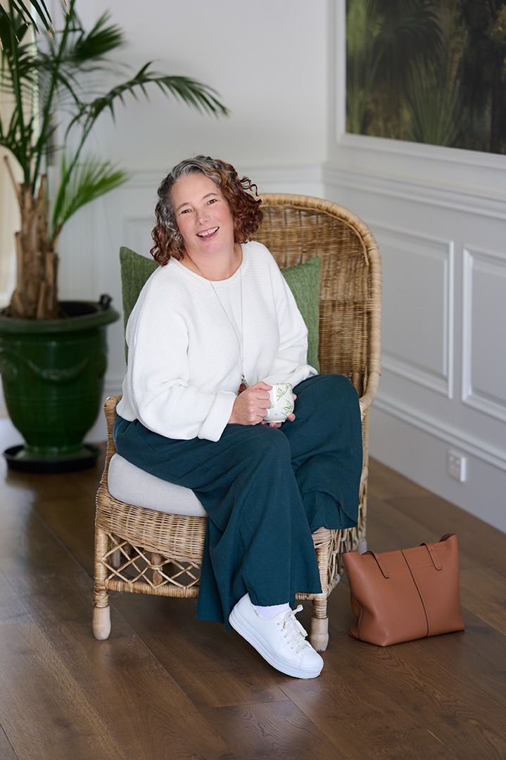A smiling woman with curly hair leans against a wall holding a floral mug, wearing jeans and a tan cardigan.