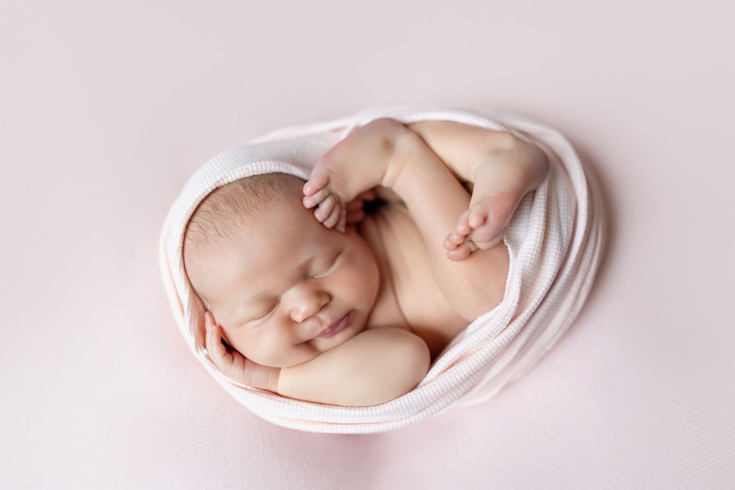 Newborn baby sleeping peacefully wrapped in white cloth, curled in fetal position on soft pink background.