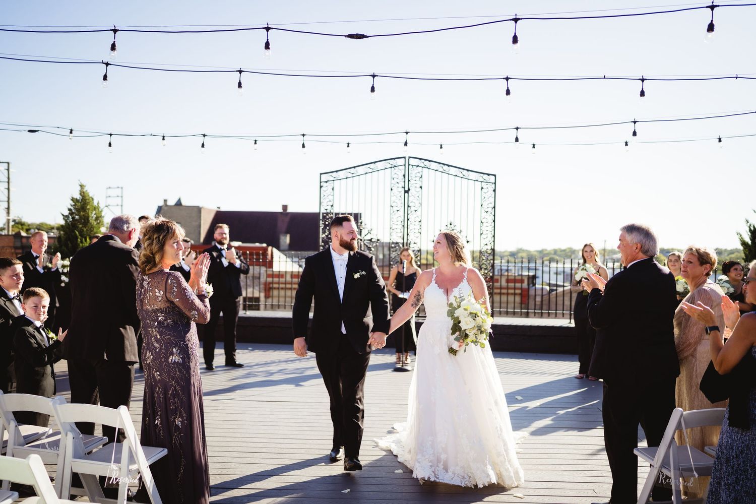 Bride and groom walk down the aisle at an outdoor rooftop wedding ceremony with string lights and guests applauding.