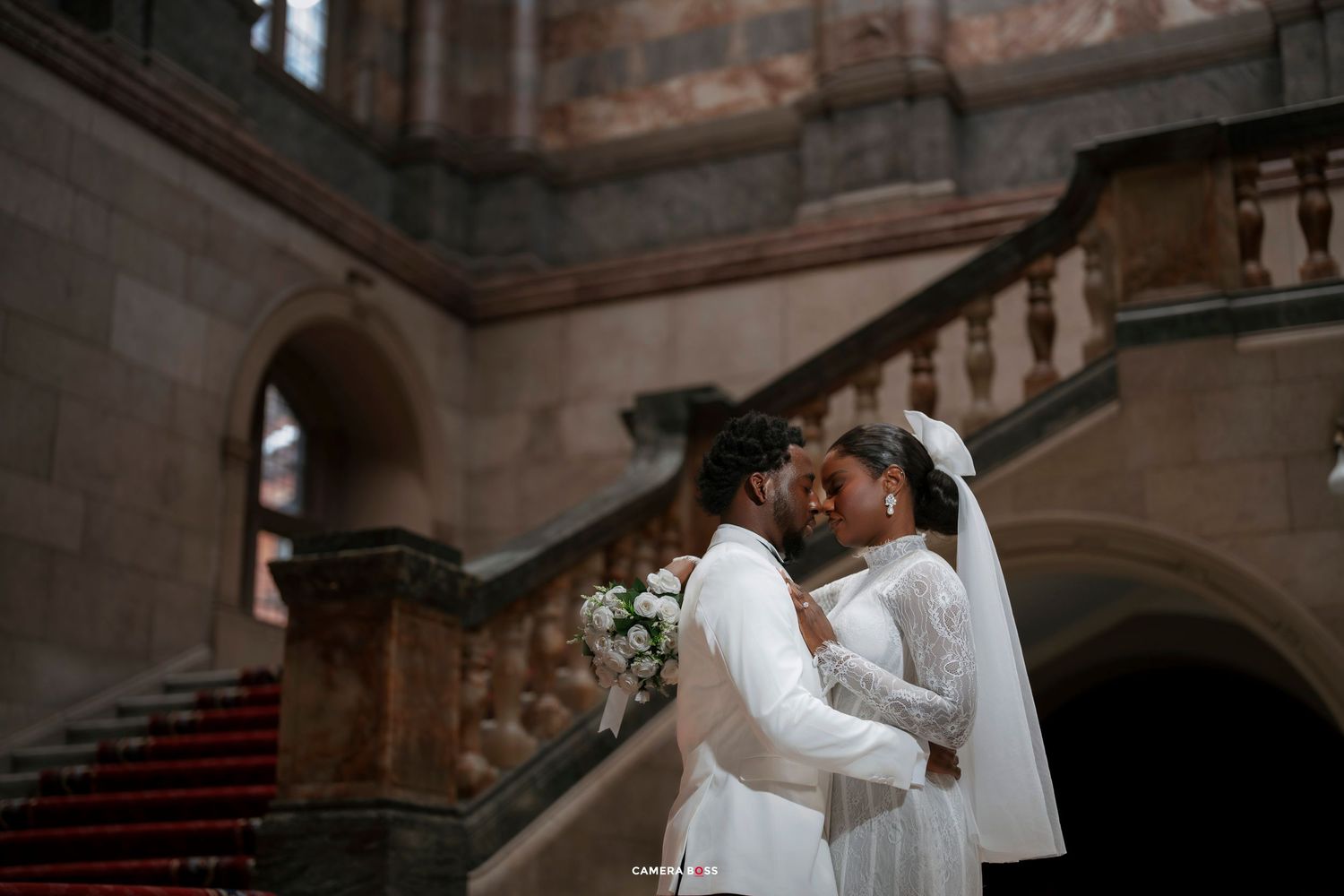 Two brides in white gowns share a romantic moment on grand staircase holding a bouquet.