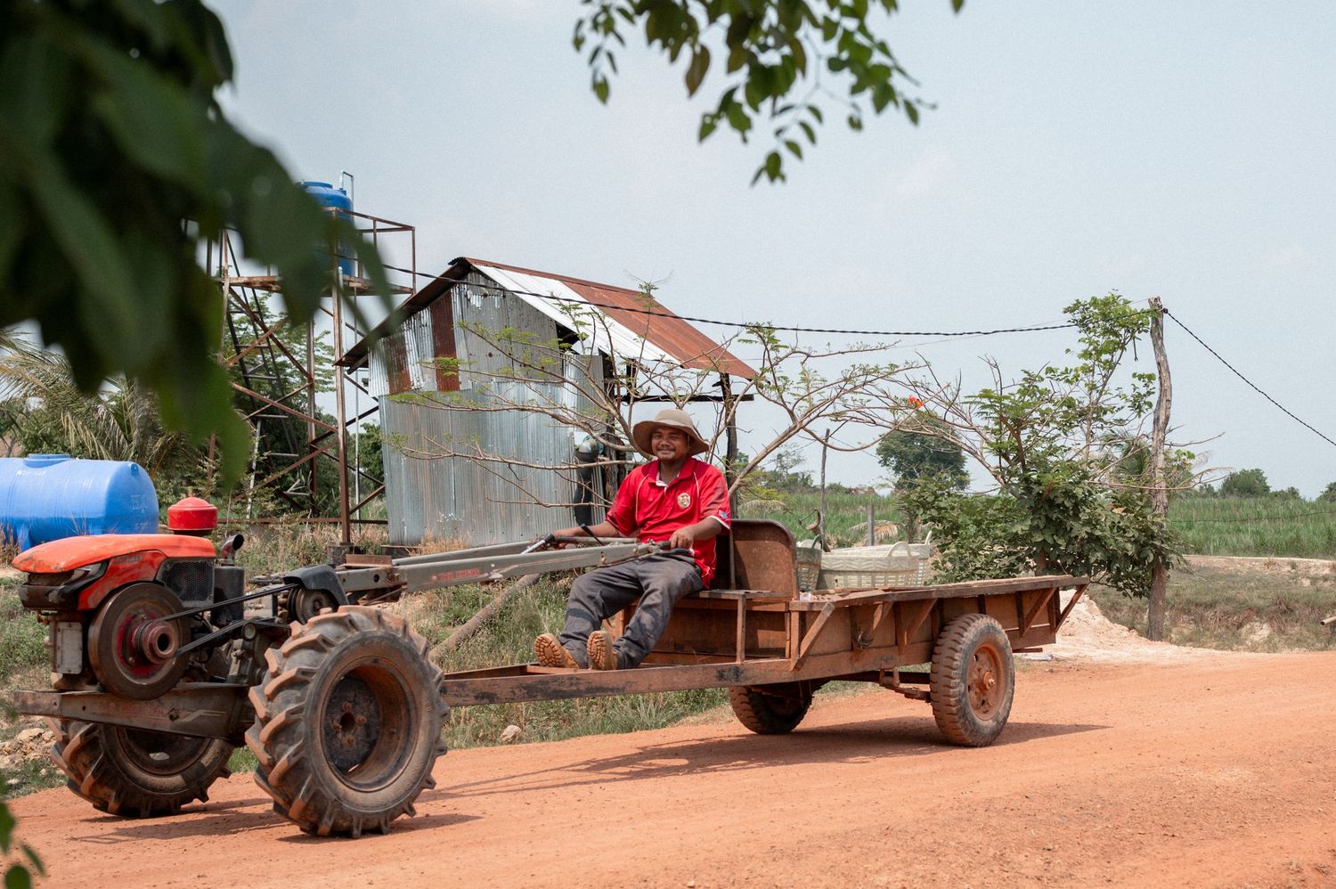 Man driving a red tractor pulling a flatbed trailer on a dusty rural dirt road in Cambodia.