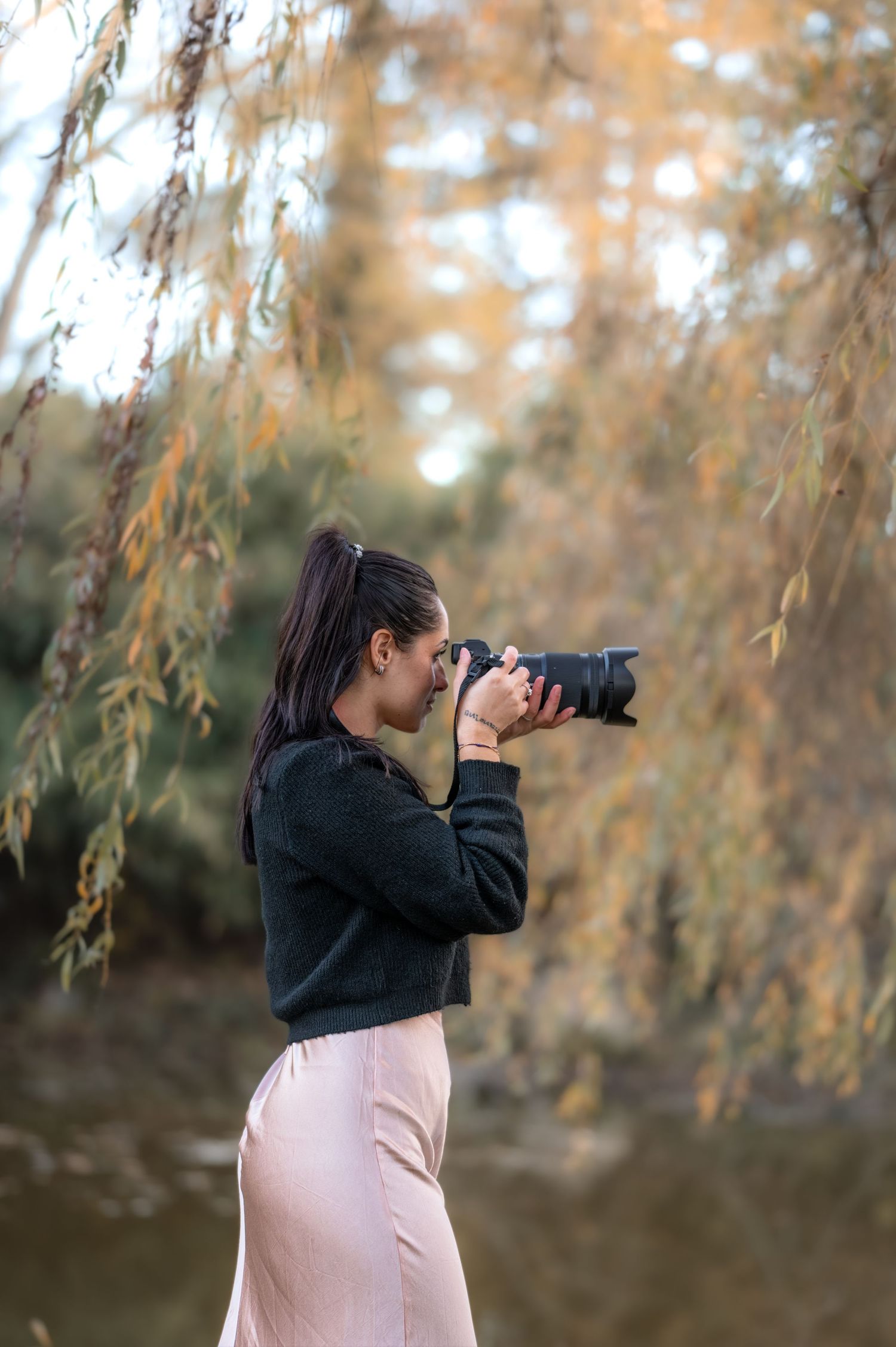 Young woman in black sweater and pink skirt photographs nature with DSLR camera under willow tree branches.