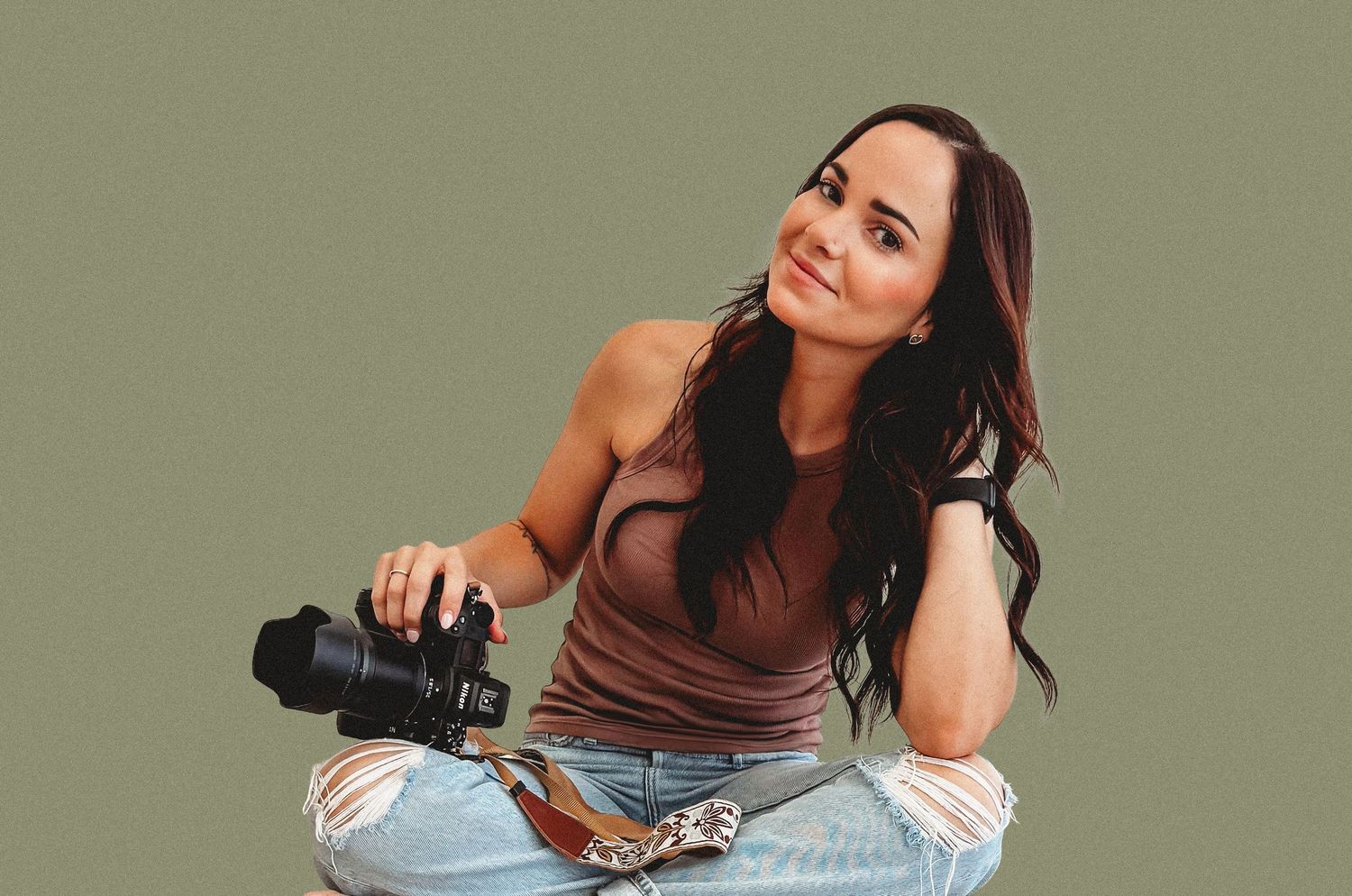 Female photographer sitting cross-legged holding camera and smiling against sage green background.
