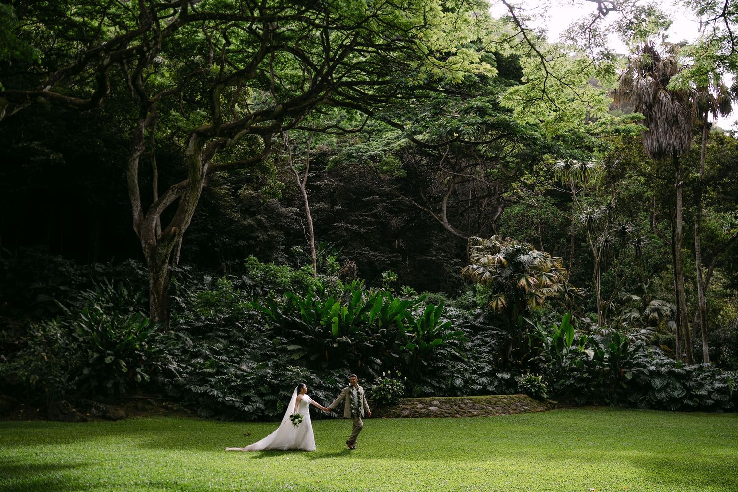 Bride and groom holding hands walking across lush green lawn surrounded by tropical gardens and rocky cliffs.
