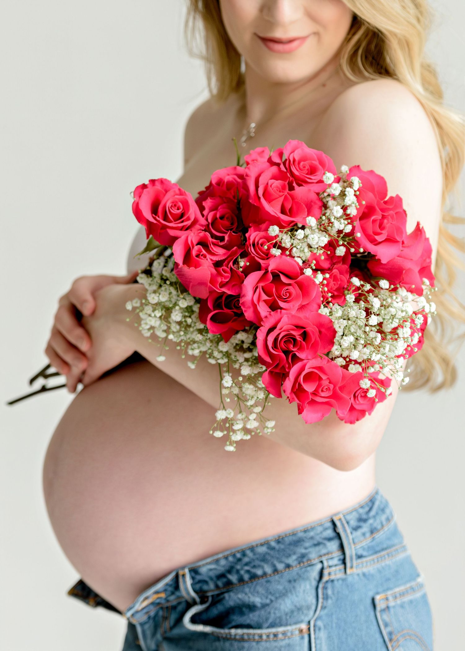 Pregnant woman in jeans holding vibrant pink roses and baby's breath bouquet during maternity photo session in Austin, TX.