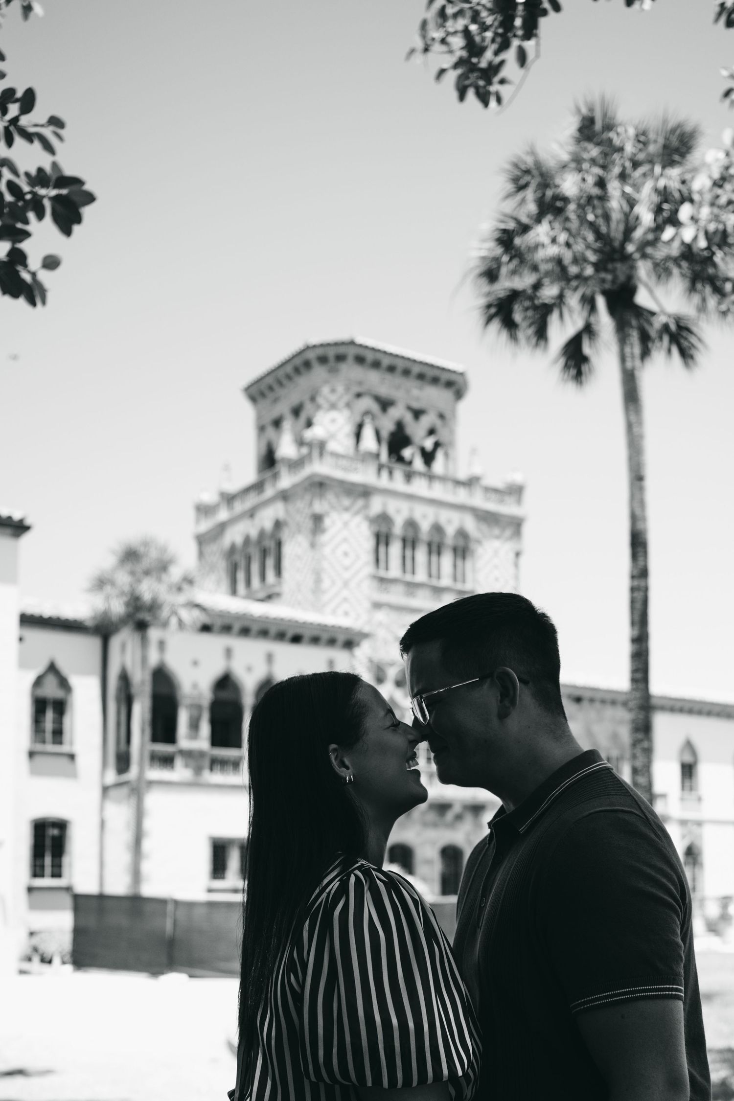Couple posing on ornate stone balustrade overlooking waterway with palm trees, man in blue polo and woman in striped dress.