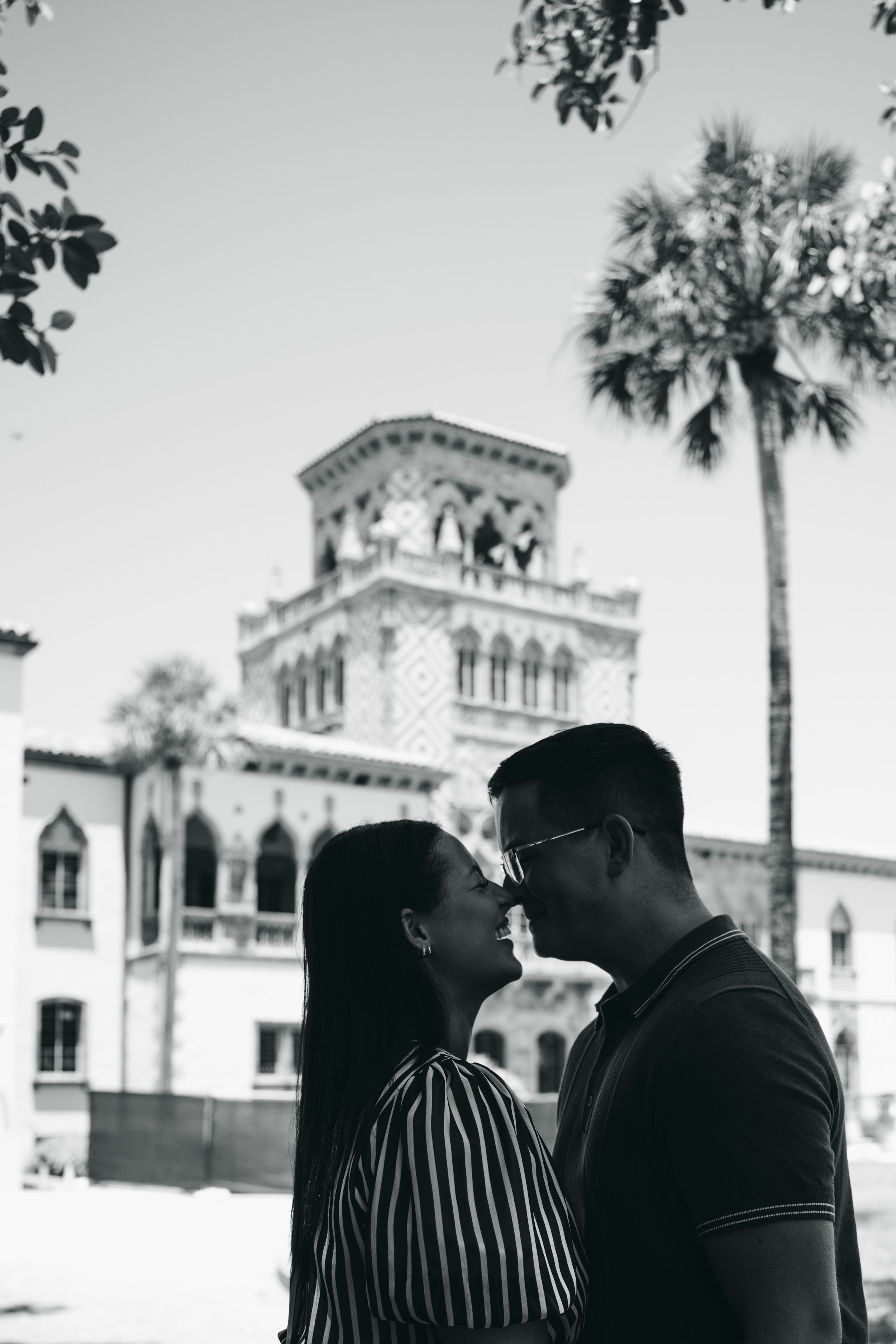 Couple posing on ornate stone balustrade overlooking waterway with palm trees, man in blue polo and woman in striped dress.