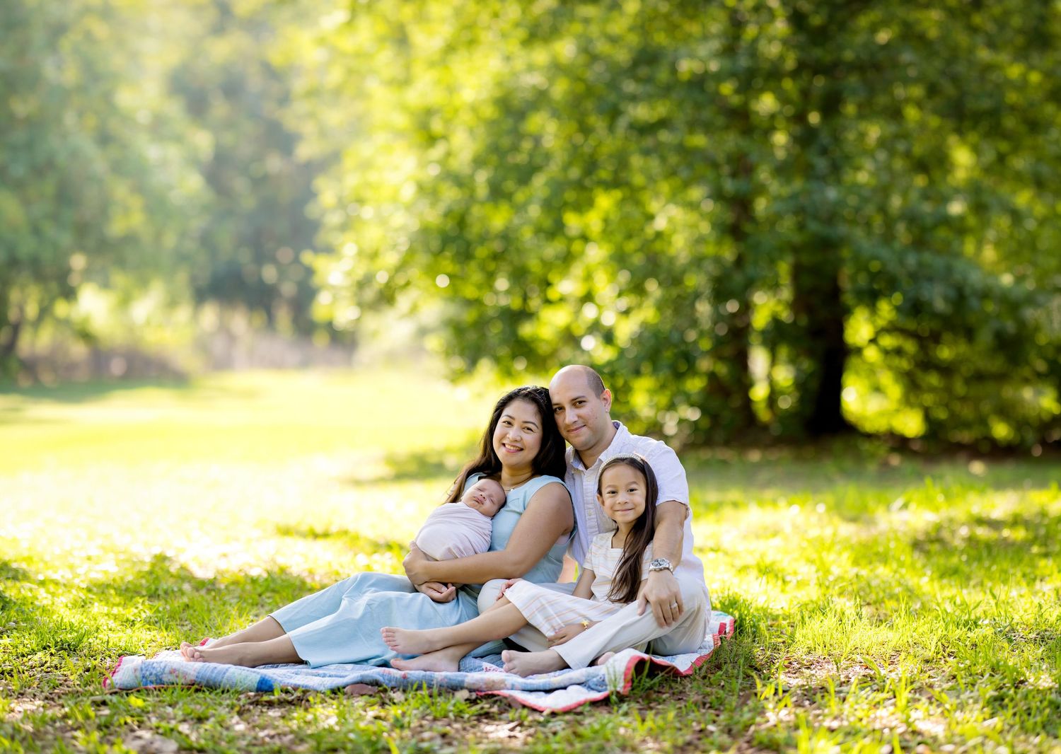 Family of four sitting on grass in sunny park, parents smiling with baby and young girl in white dresses.