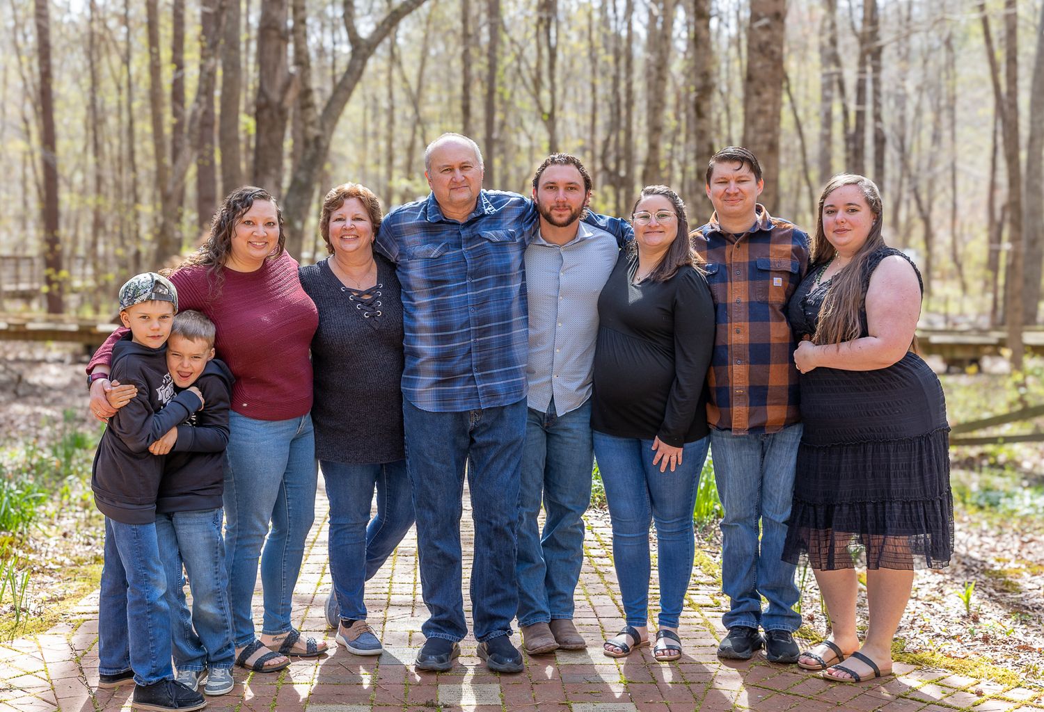 Large family group portrait outdoors in a wooded park setting, posing together on a wooden bridge in spring.