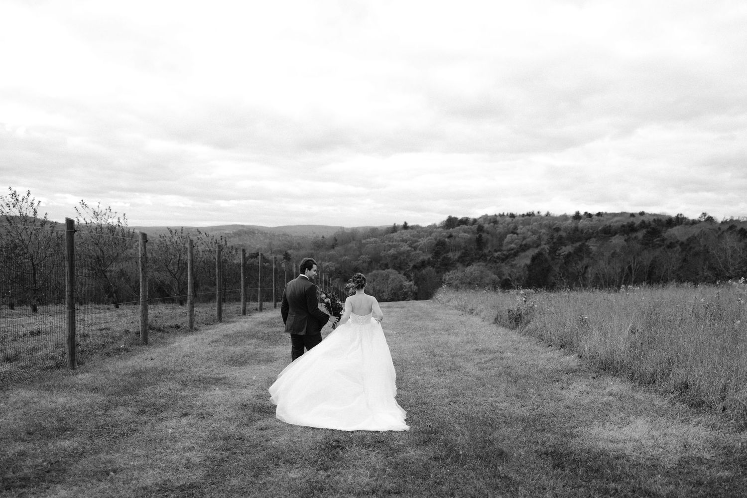 Outdoor wedding ceremony at a rustic red barn venue with guests gathered beneath a draped arch on a cloudy day.